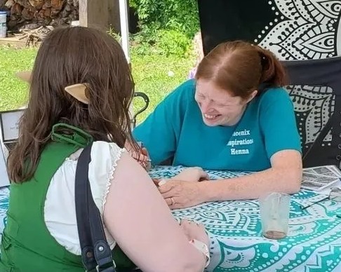 Two women sitting at a table, smiling and engaging in conversation outdoors, with greenery in the background.