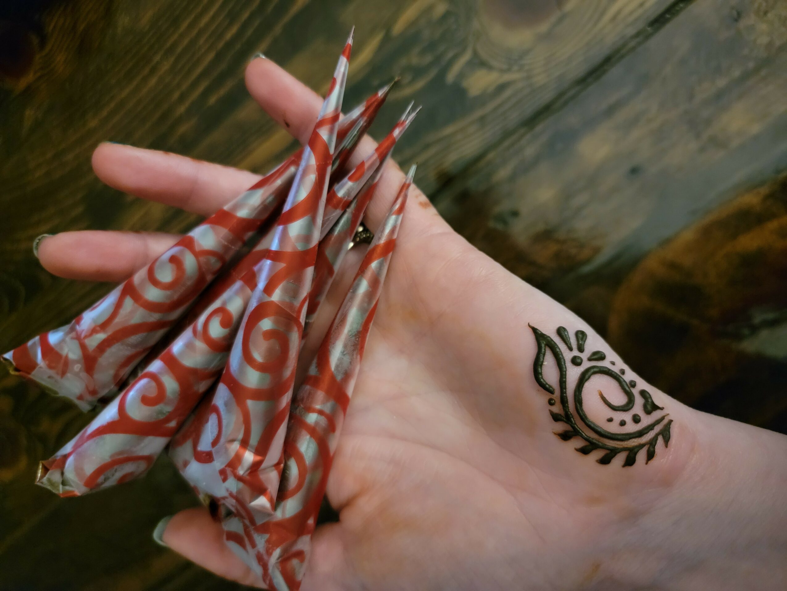 Hand with a black tattoo holding a colorful fabric with red, white, and orange swirls, against a wooden background.