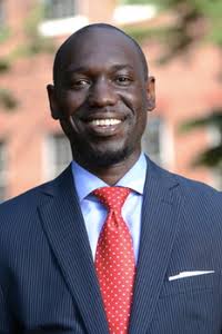 Man in a suit and red tie smiling outdoors with blurred background