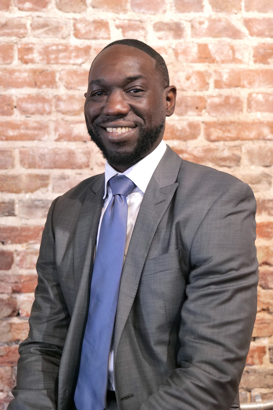 Man in gray suit and blue tie smiling in front of brick wall, facing forward.