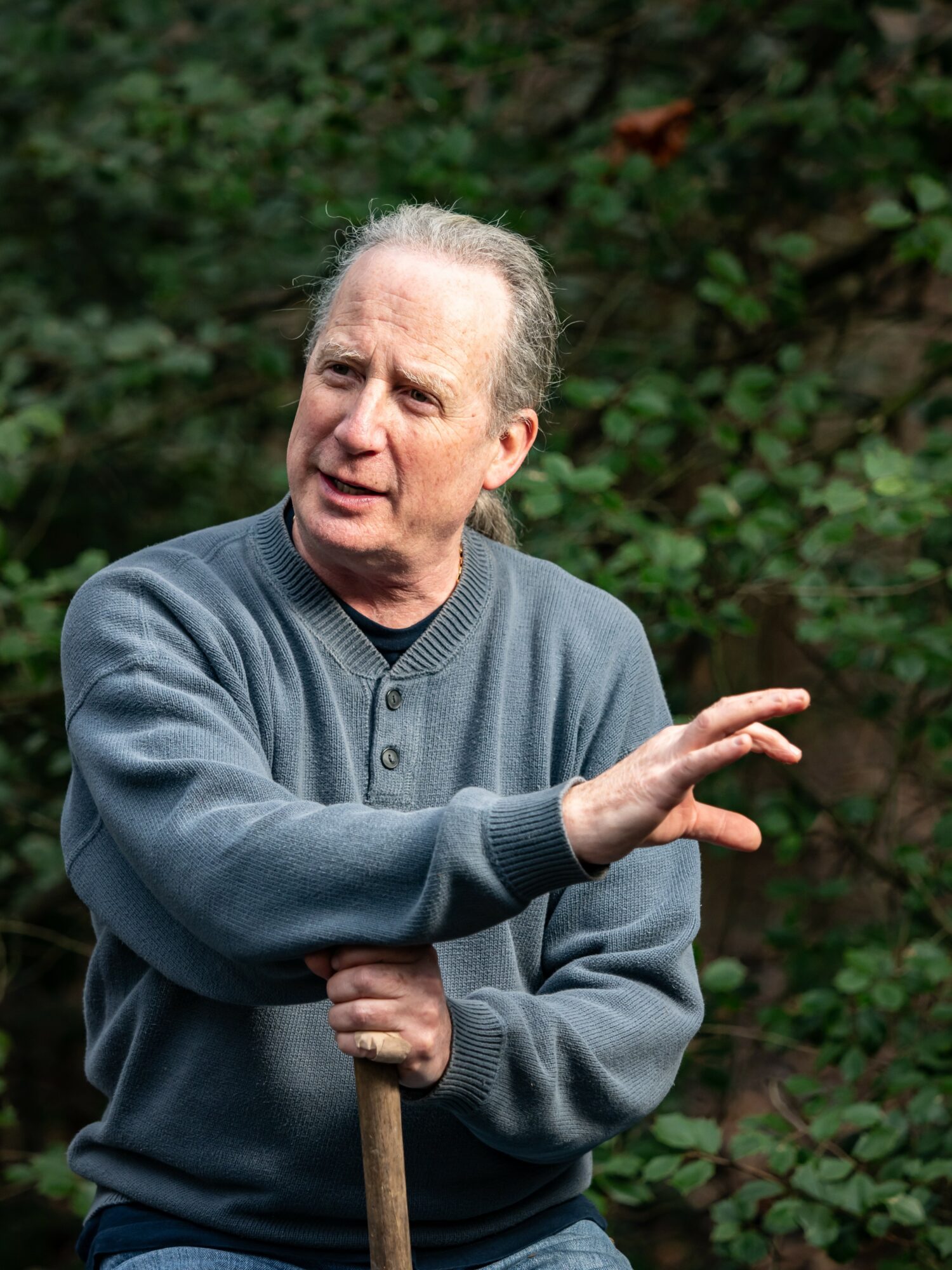 Older man with gray hair in a gray sweater gesturing with his right hand outdoors, green foliage background.