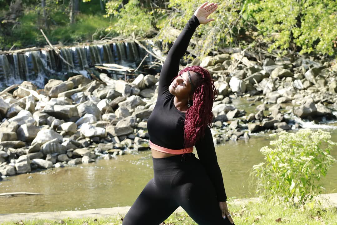 Woman with long red hair stretching outdoors near a river with rocks and greenery.