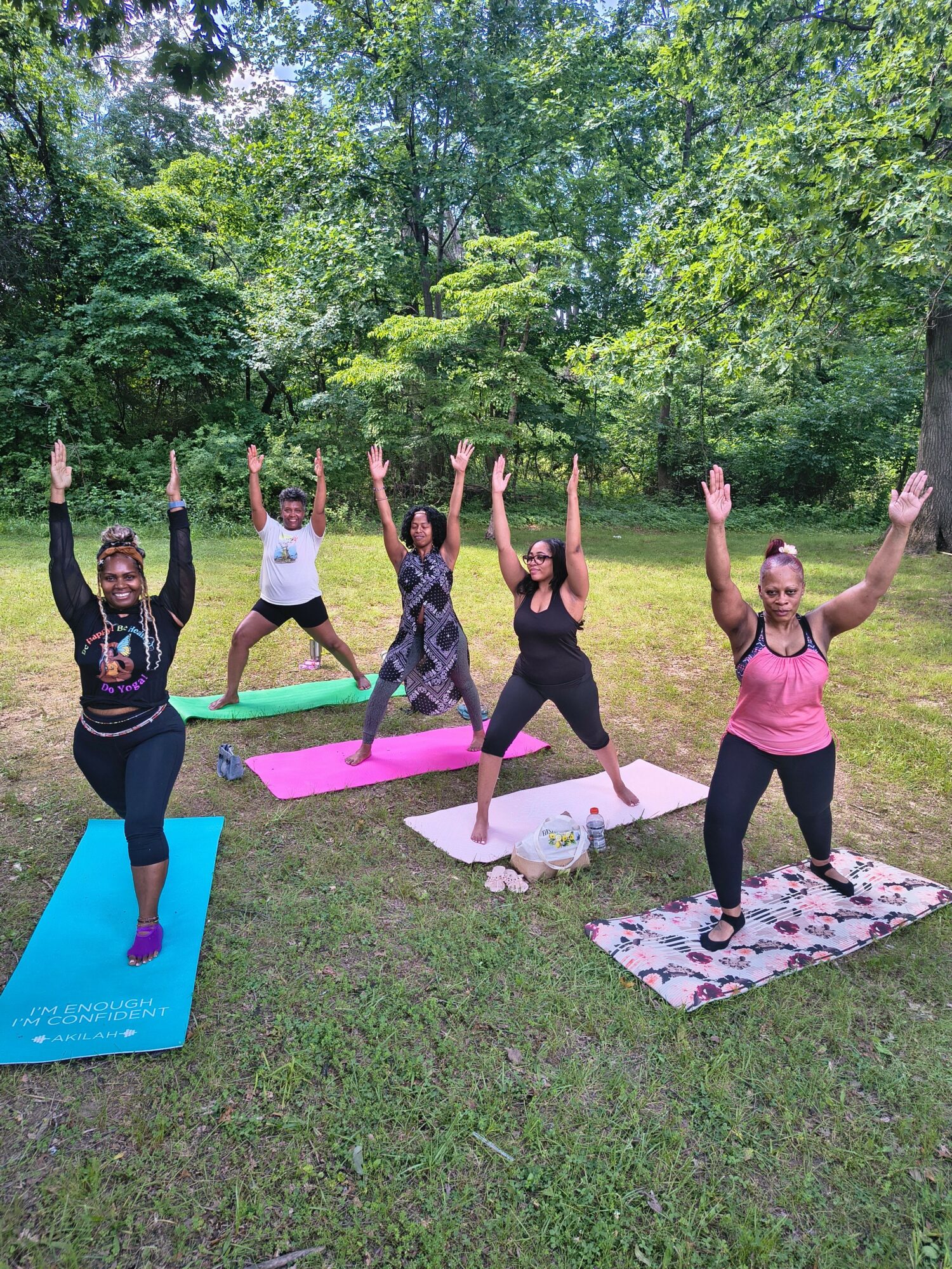 Group of six people practicing yoga outdoors on mats in a grassy area with trees, in various poses.