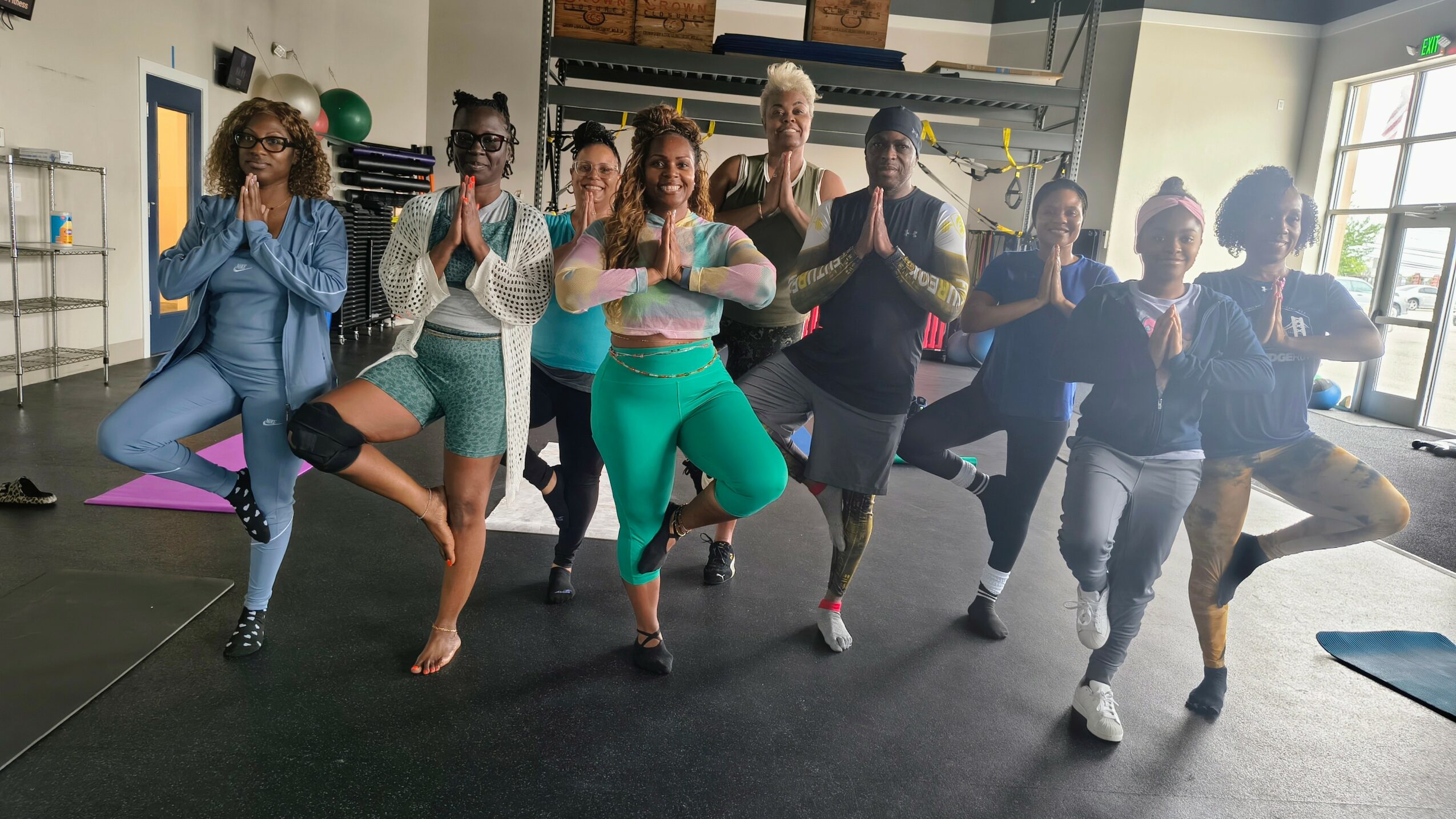 Group of people practicing yoga in a studio, standing on one leg with hands in prayer position, smiling.