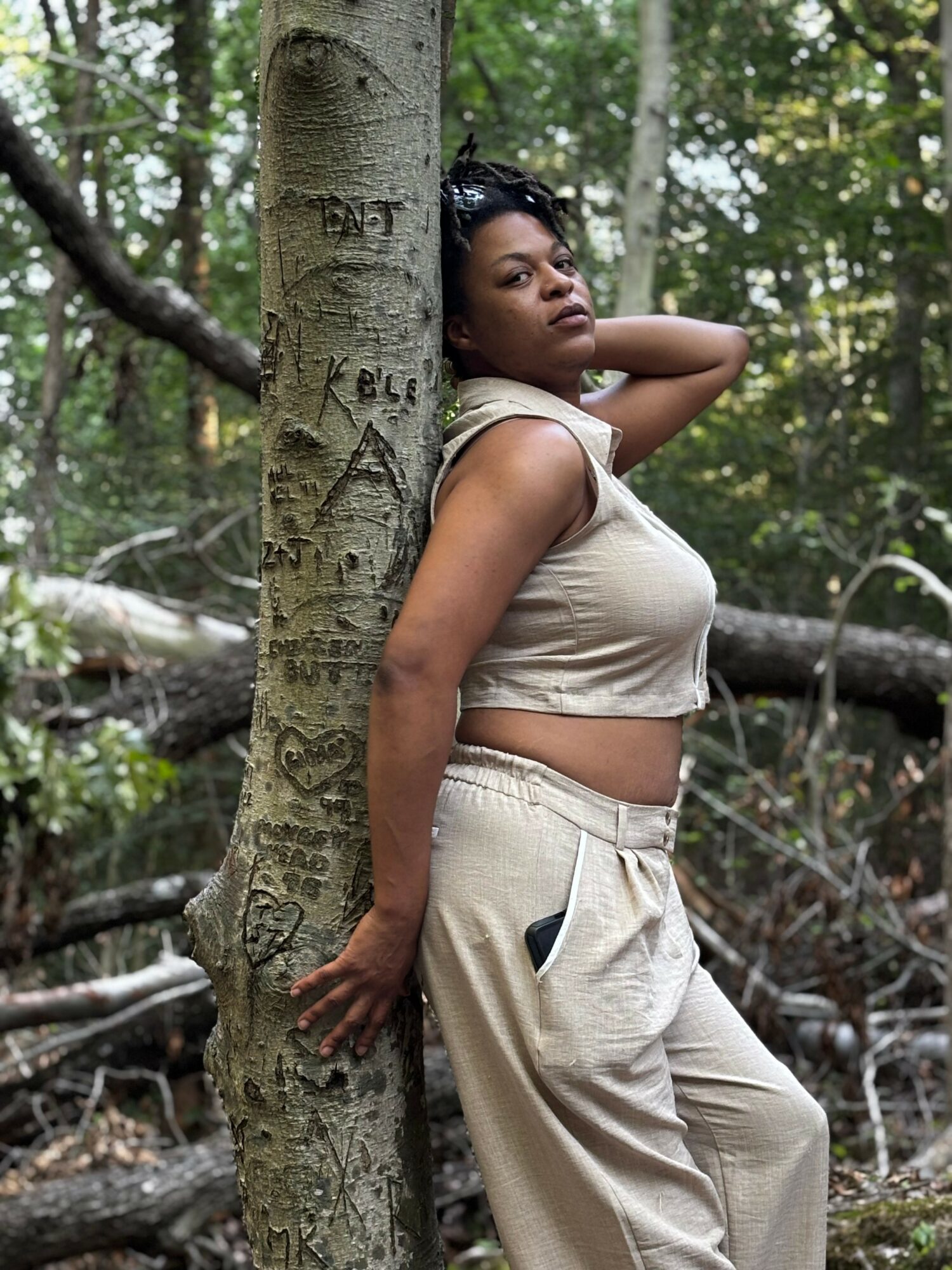 Woman leaning against a tree in a forest, looking into the distance, wearing a beige crop top and pants.