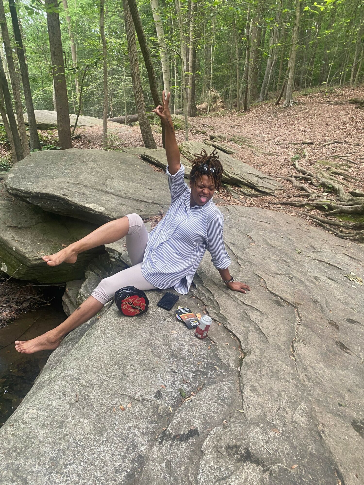 Person sitting on a large rock in a forest, holding a tree branch, with a backpack and water bottle nearby.
