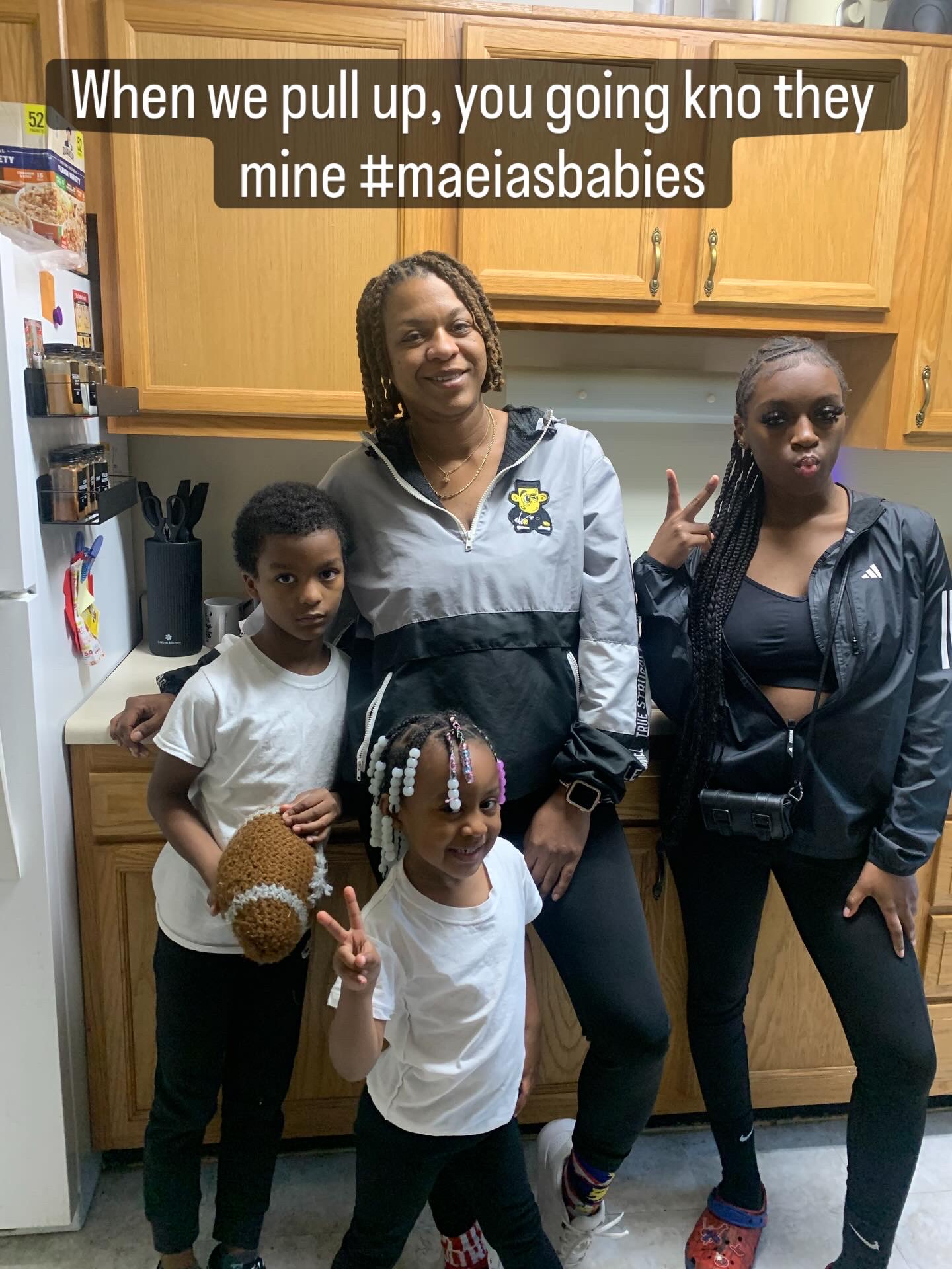Three women and two children in a kitchen, one woman smiling, children holding a teddy bear and making peace signs.
