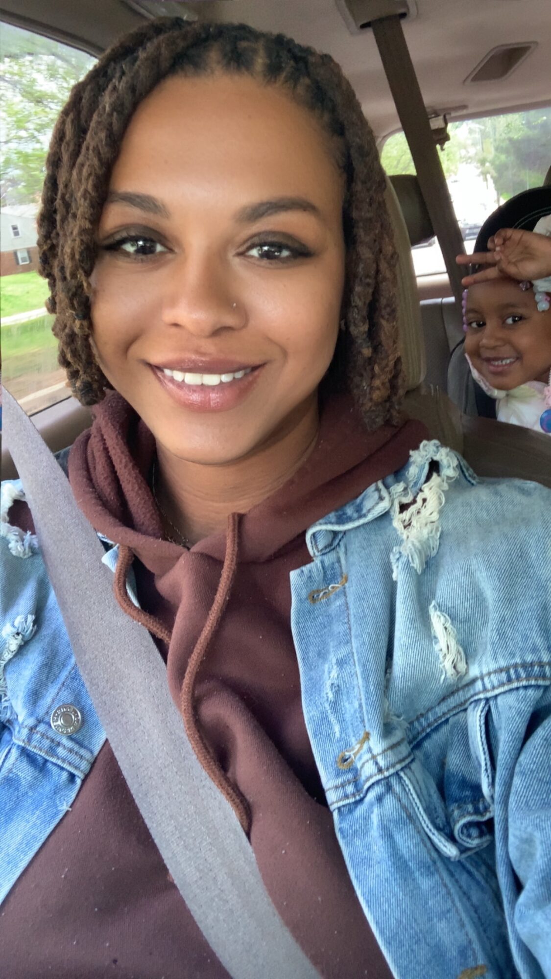 Woman smiling in car with child in background, wearing denim jacket and brown hoodie, seatbelt visible.