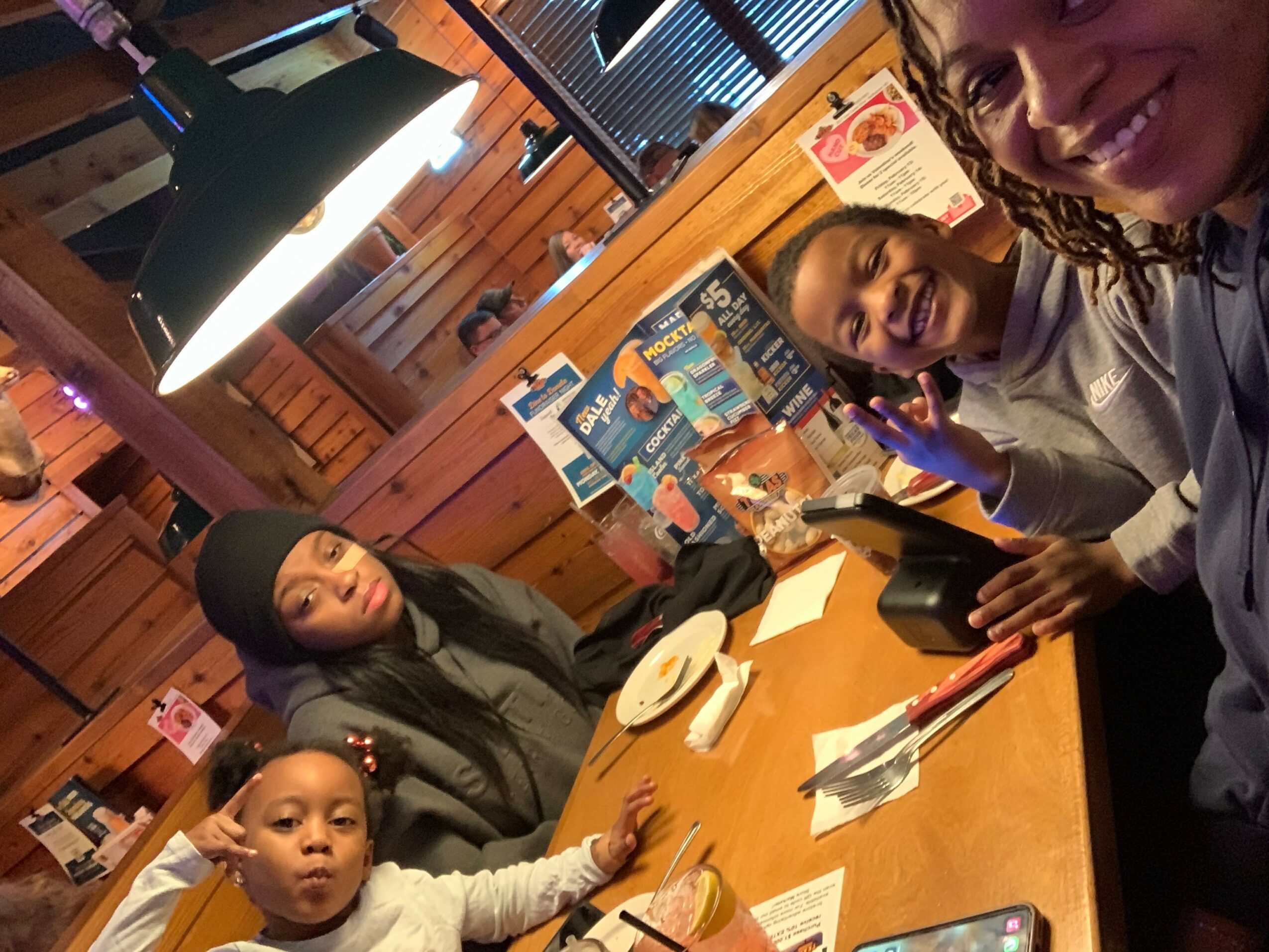 Four people sitting at a restaurant table smiling, with food and drinks, and a woman taking a selfie.