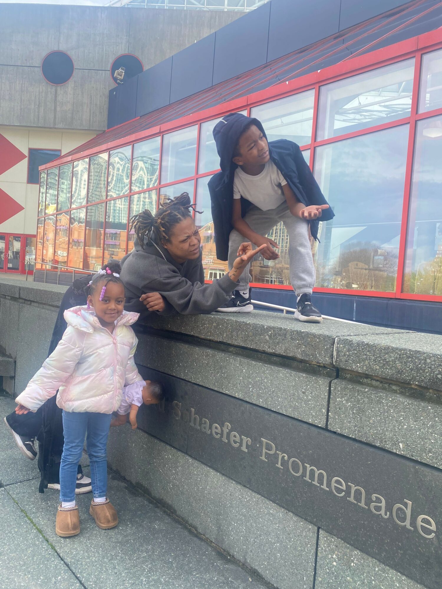 Two children and an adult standing outside a building with red window frames, one child in a pink jacket, the other in a gray jacket.
