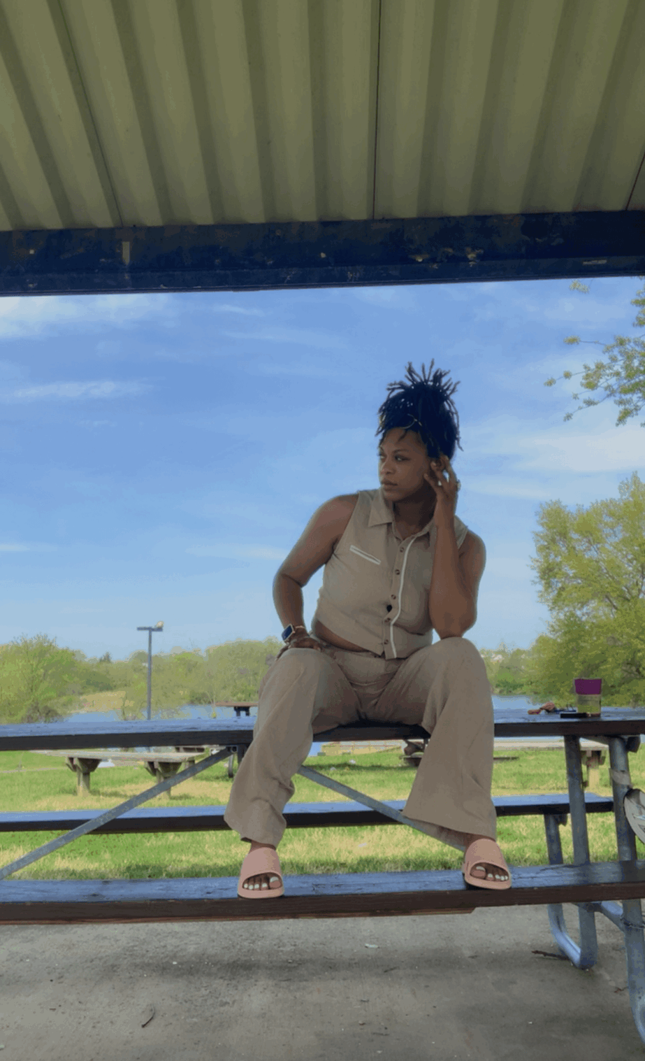 Person sitting on a picnic table under a shelter, with trees and blue sky in the background.