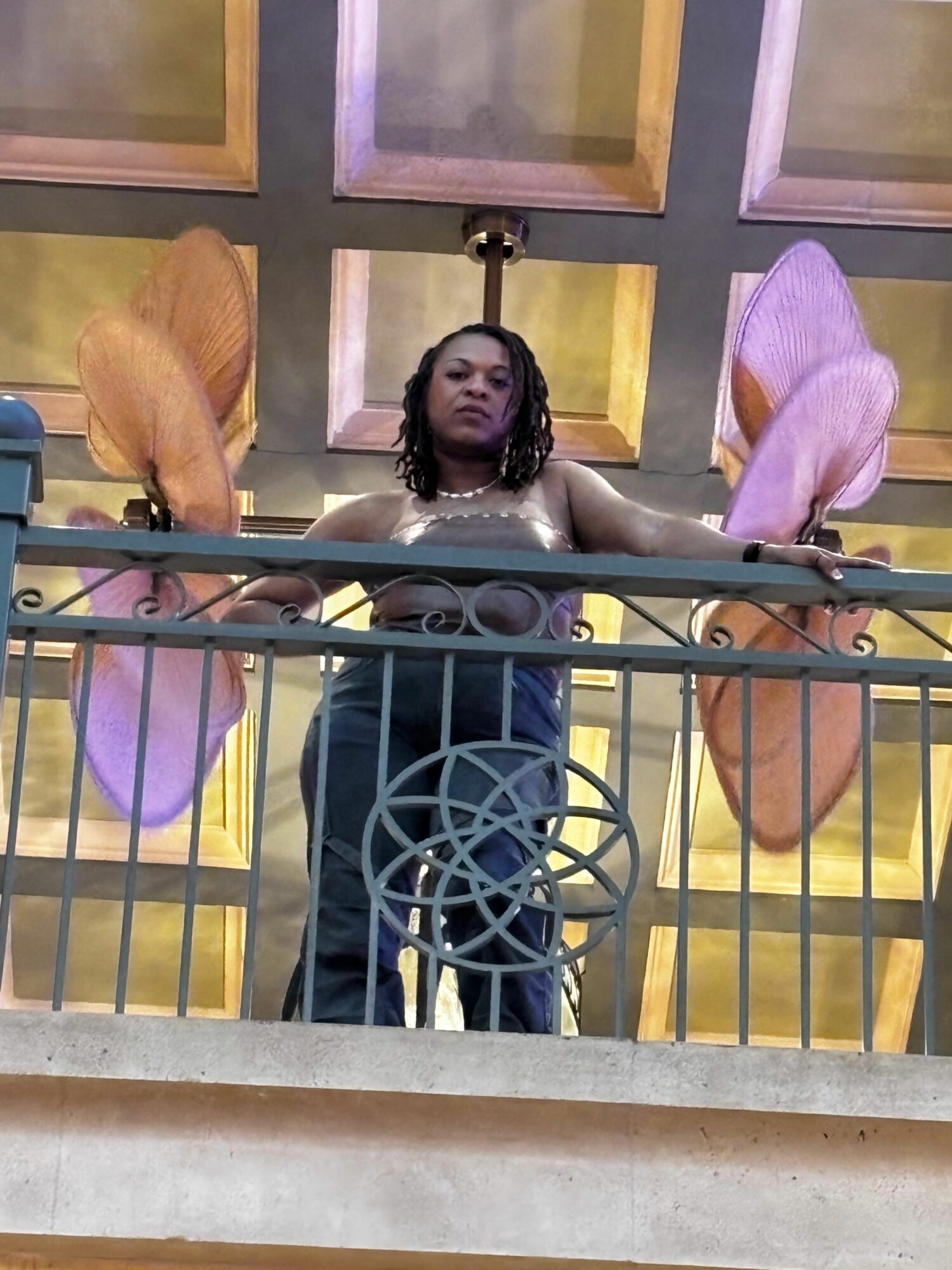 Person with fairy wings leaning on a railing, looking at the camera, with colorful wings and a dark outfit.