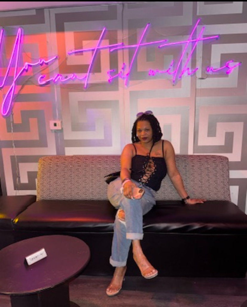 Woman sitting on a sofa in front of a geometric patterned wall with neon pink and purple sign, wearing a black top and jeans.