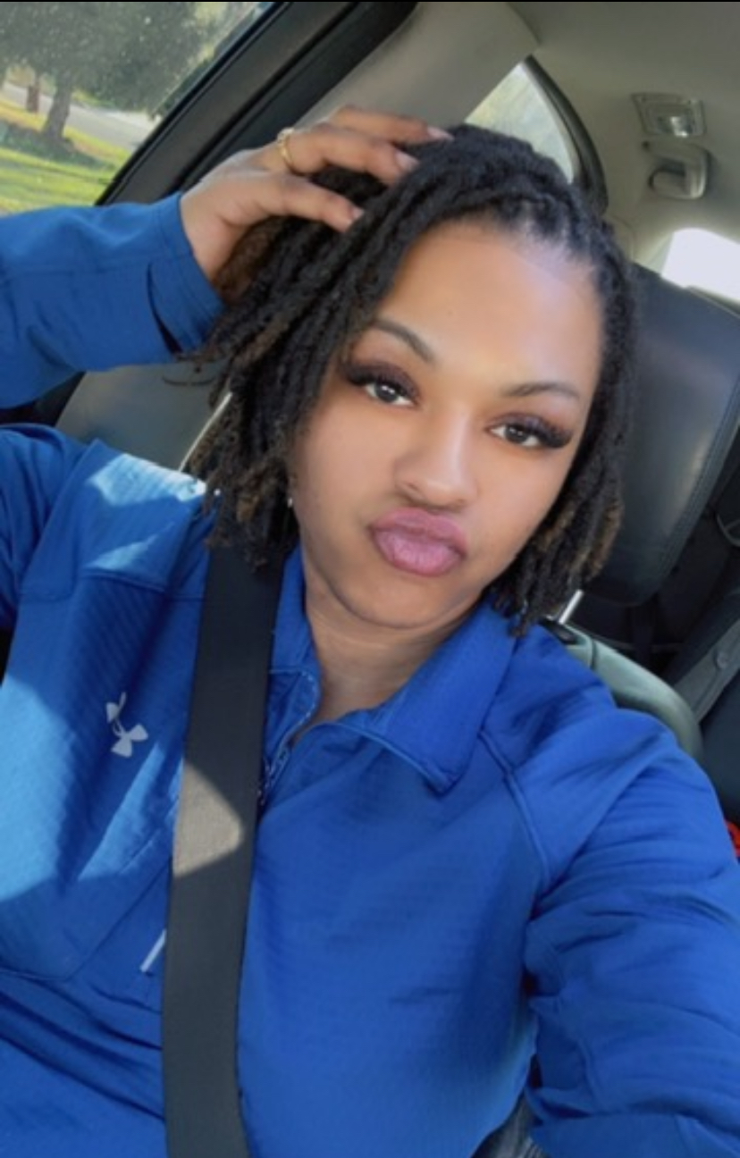 Woman with short dreadlocks taking a selfie inside a car, wearing a blue jacket, making a pouty face.