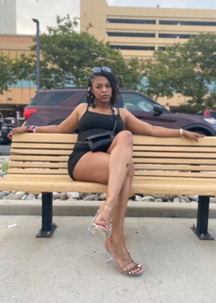 Woman sitting on a bench with arms stretched out, wearing a black outfit and clear high heels, in an outdoor urban setting.