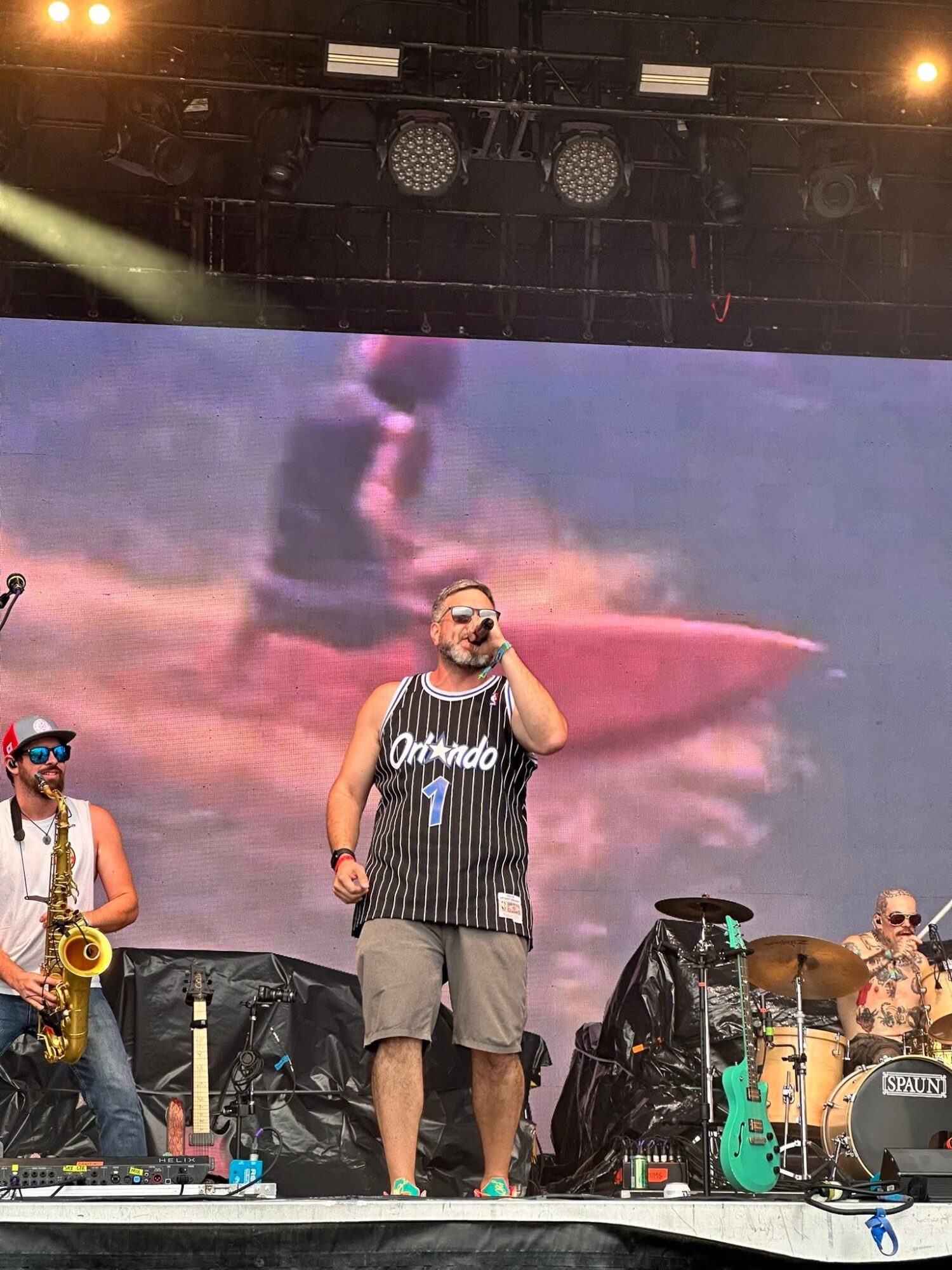 Man in a black Orlando basketball jersey and shorts singing on stage with musicians, large screen behind showing a boat and water.