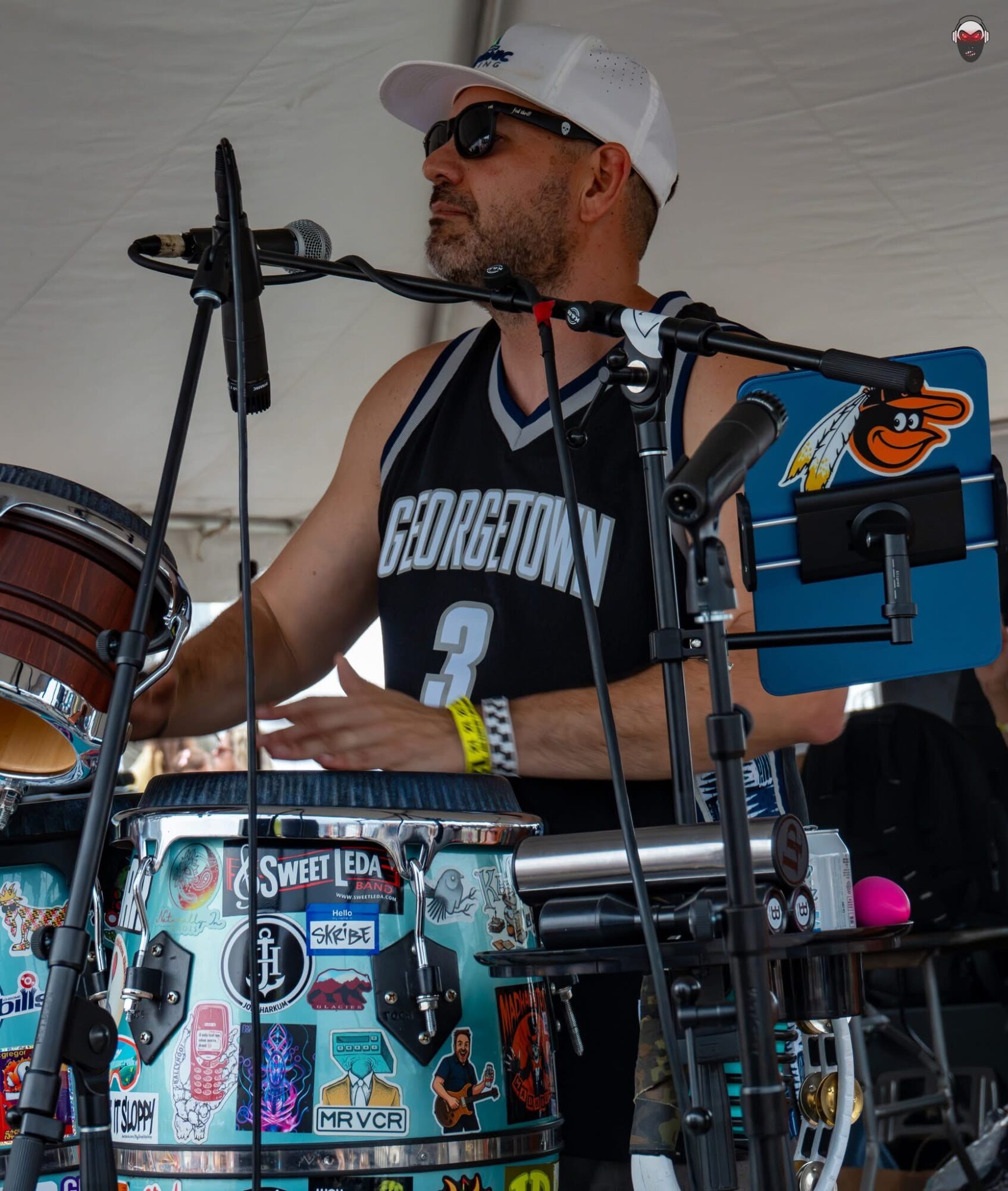 Man wearing sunglasses, a white cap, and a Georgetown basketball jersey playing drums at an event, with a blue cooler nearby.