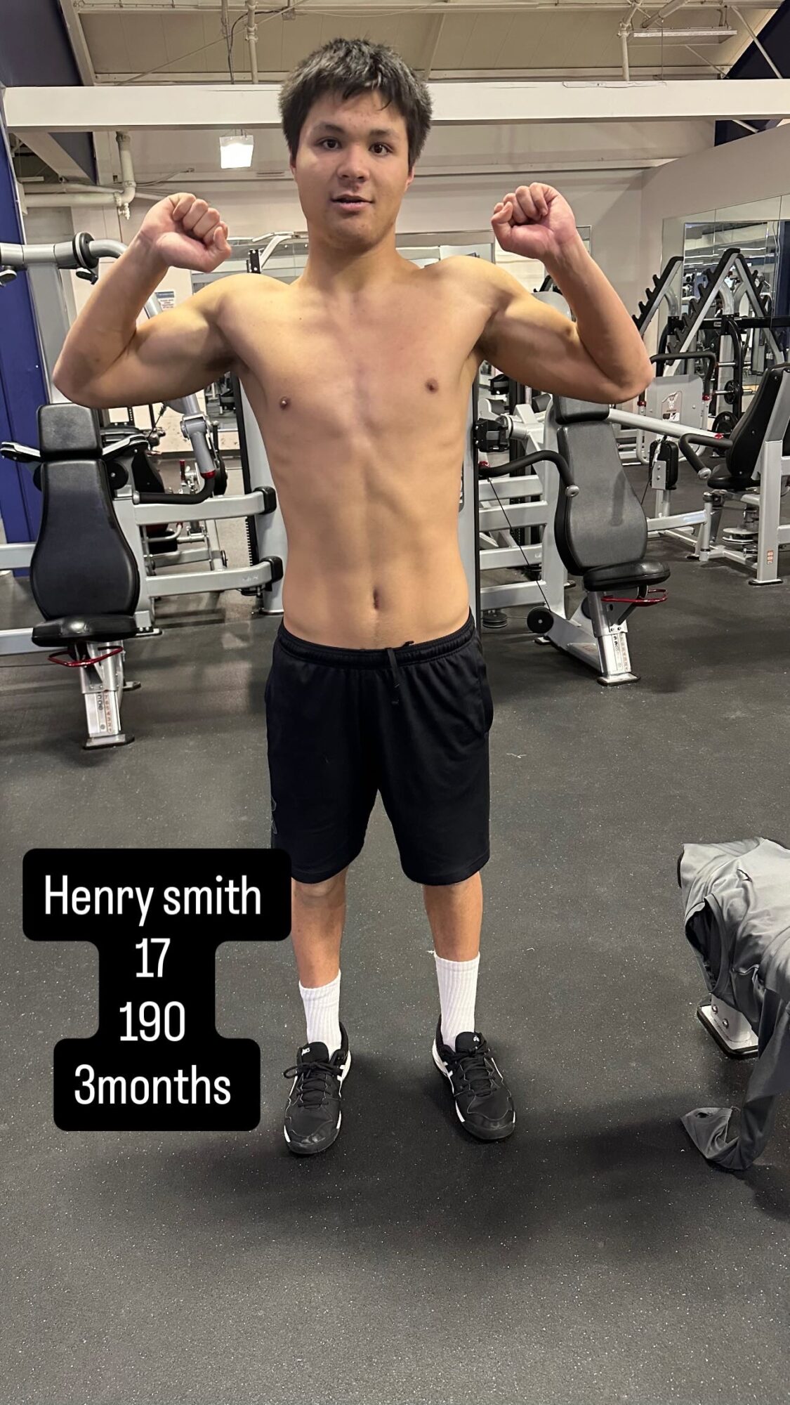 Young man with short hair, shirtless, flexing arms in gym, wearing black shorts and sneakers, gym equipment in background.
