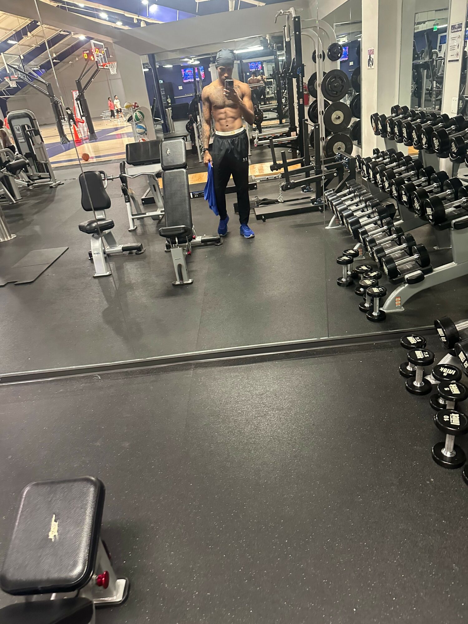 Person in gym with dumbbells and workout equipment, facing left, in a well-lit fitness center.