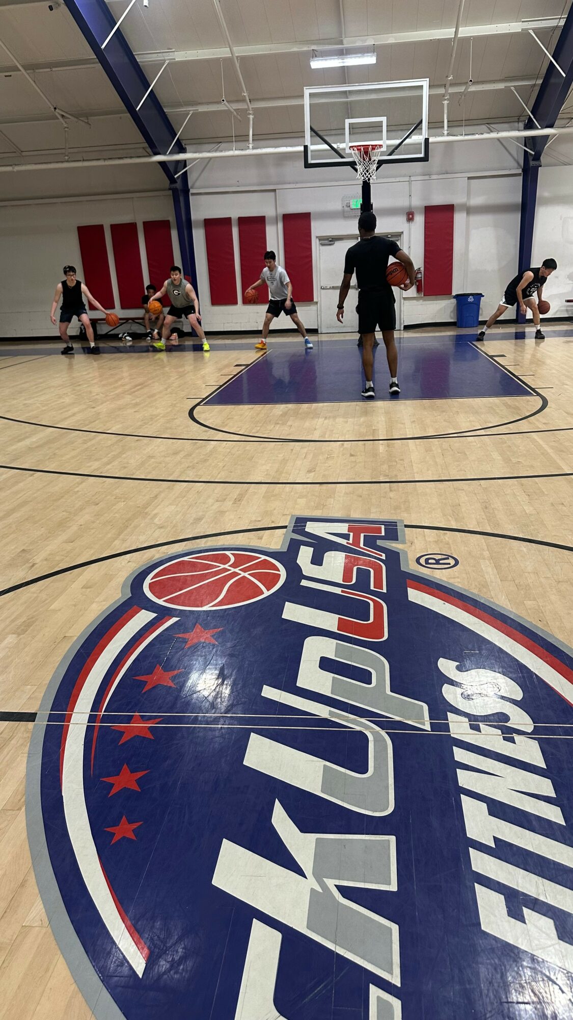 Basketball court with players practicing, and a large logo on the floor reading 'KLAYUP FITNESS'.
