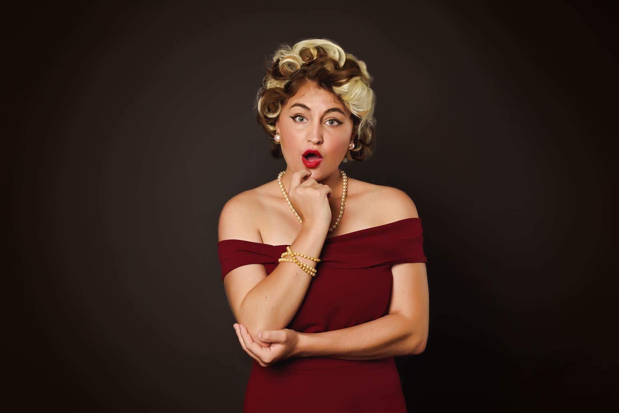 Woman with vintage hairstyle and red off-shoulder dress, posing with surprised expression against dark background.