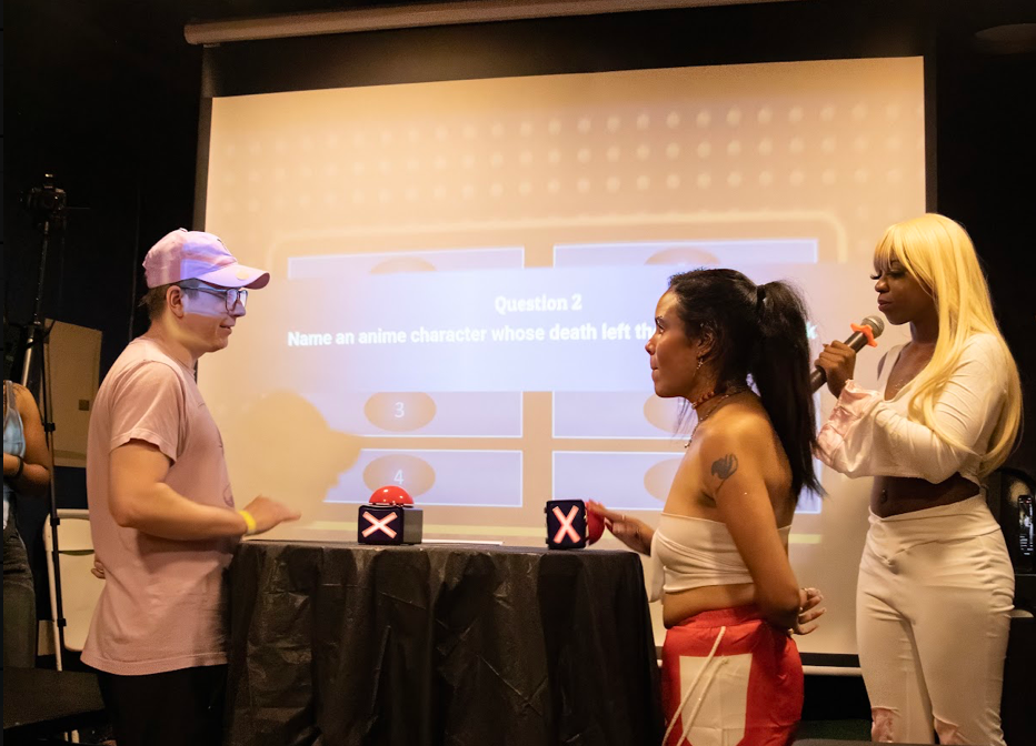 Three women stand on stage with a large screen behind them, participating in a game or quiz show, with two boxes on a table.