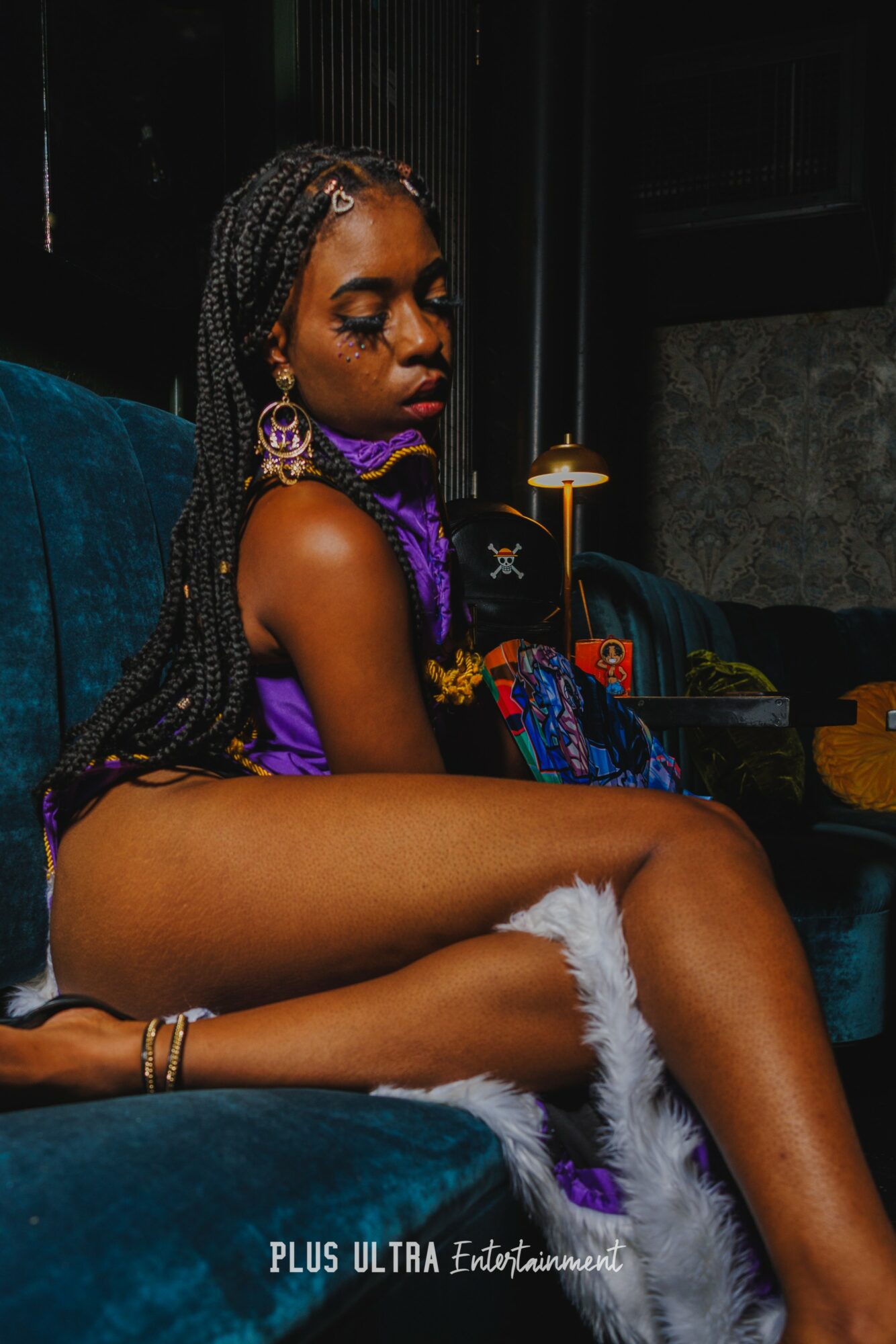 Young woman with braided hair and large earrings sitting on a blue couch, wearing colorful clothing and white furry footwear.