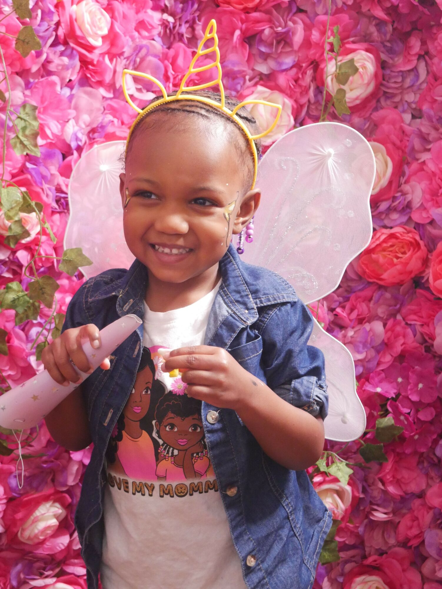 Child with a unicorn headband smiling, holding a toy, surrounded by pink and purple flowers.