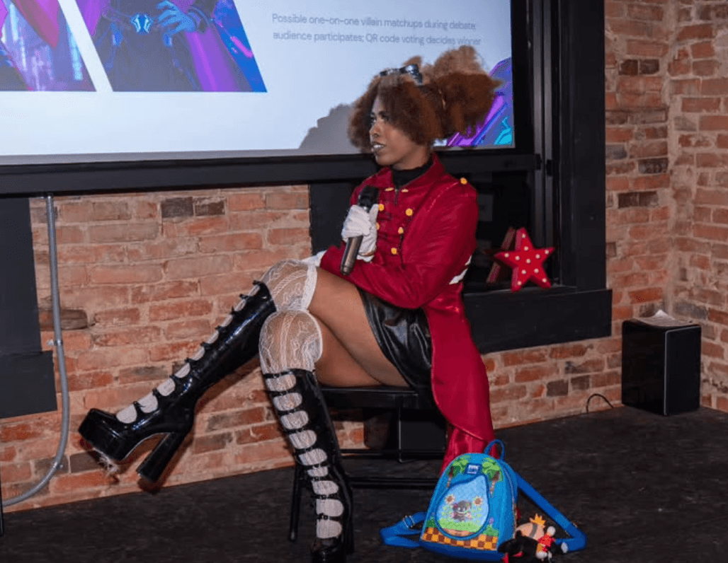 Person with curly hair in a red coat and black boots sitting on a chair against a brick wall, with a backpack on the floor.