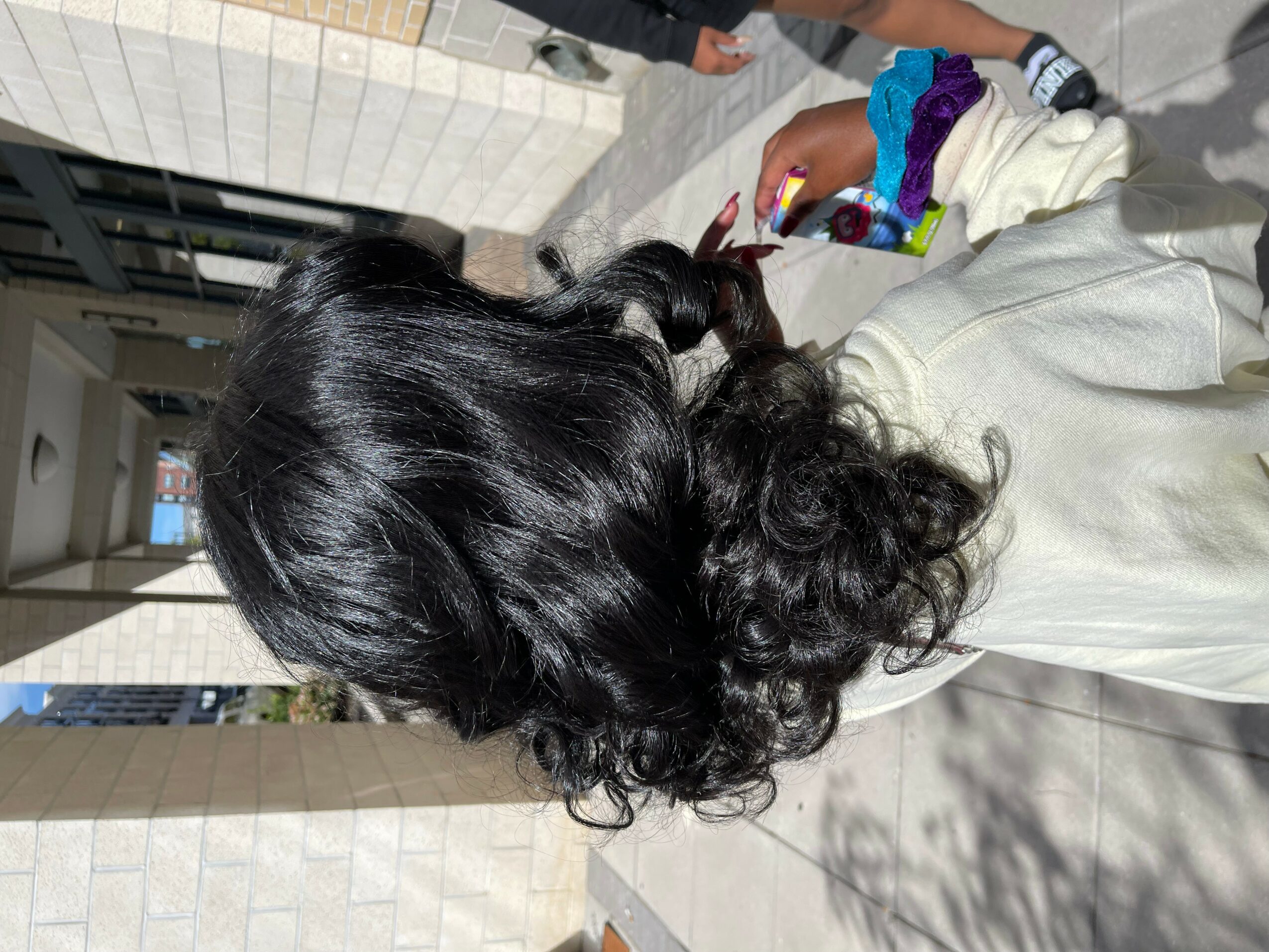 Person with long, curly black hair seen from behind, walking outdoors on a sunny day.