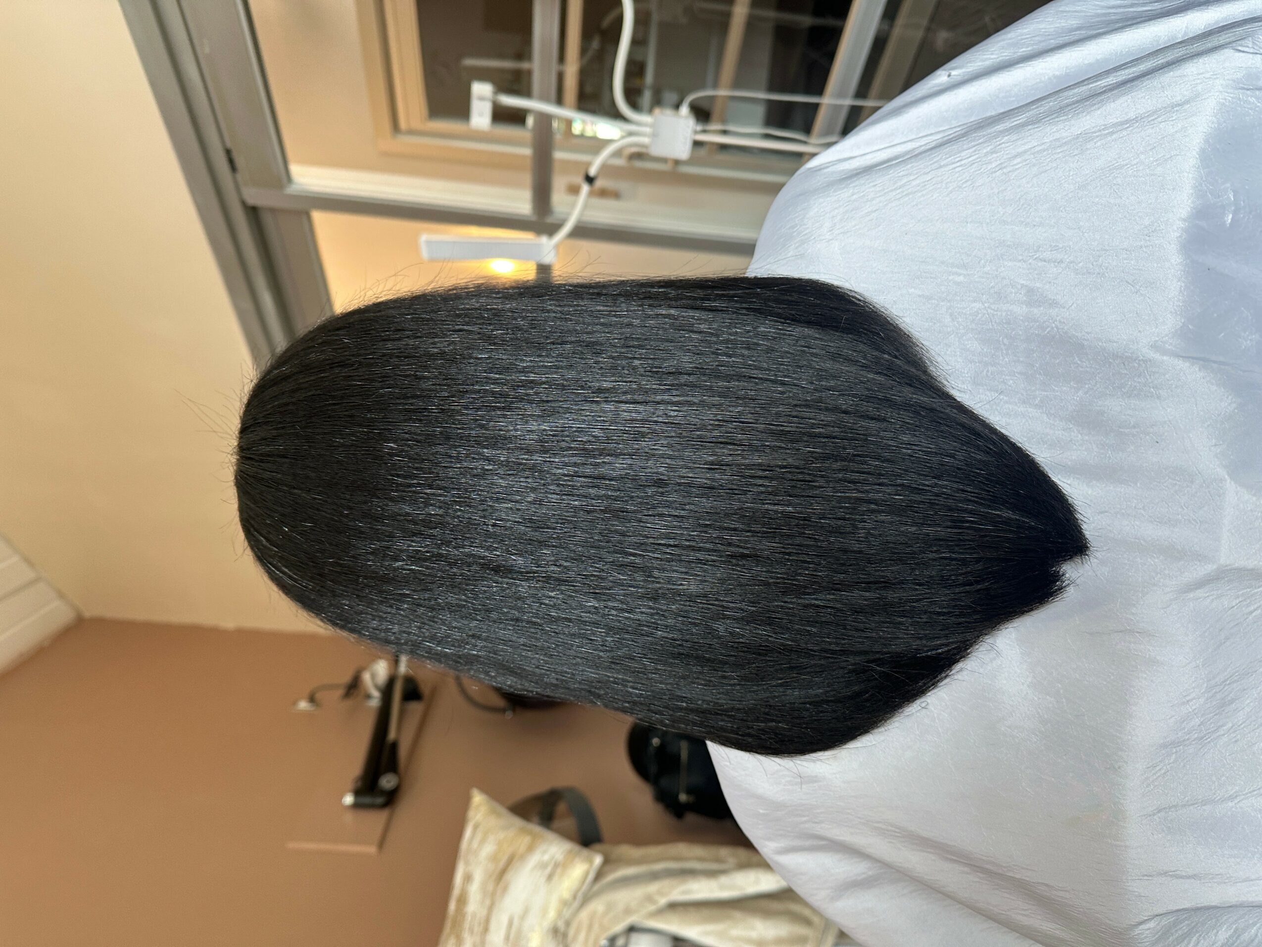 Person with straight, black hair in a salon chair, facing away, with a window and salon equipment in the background.