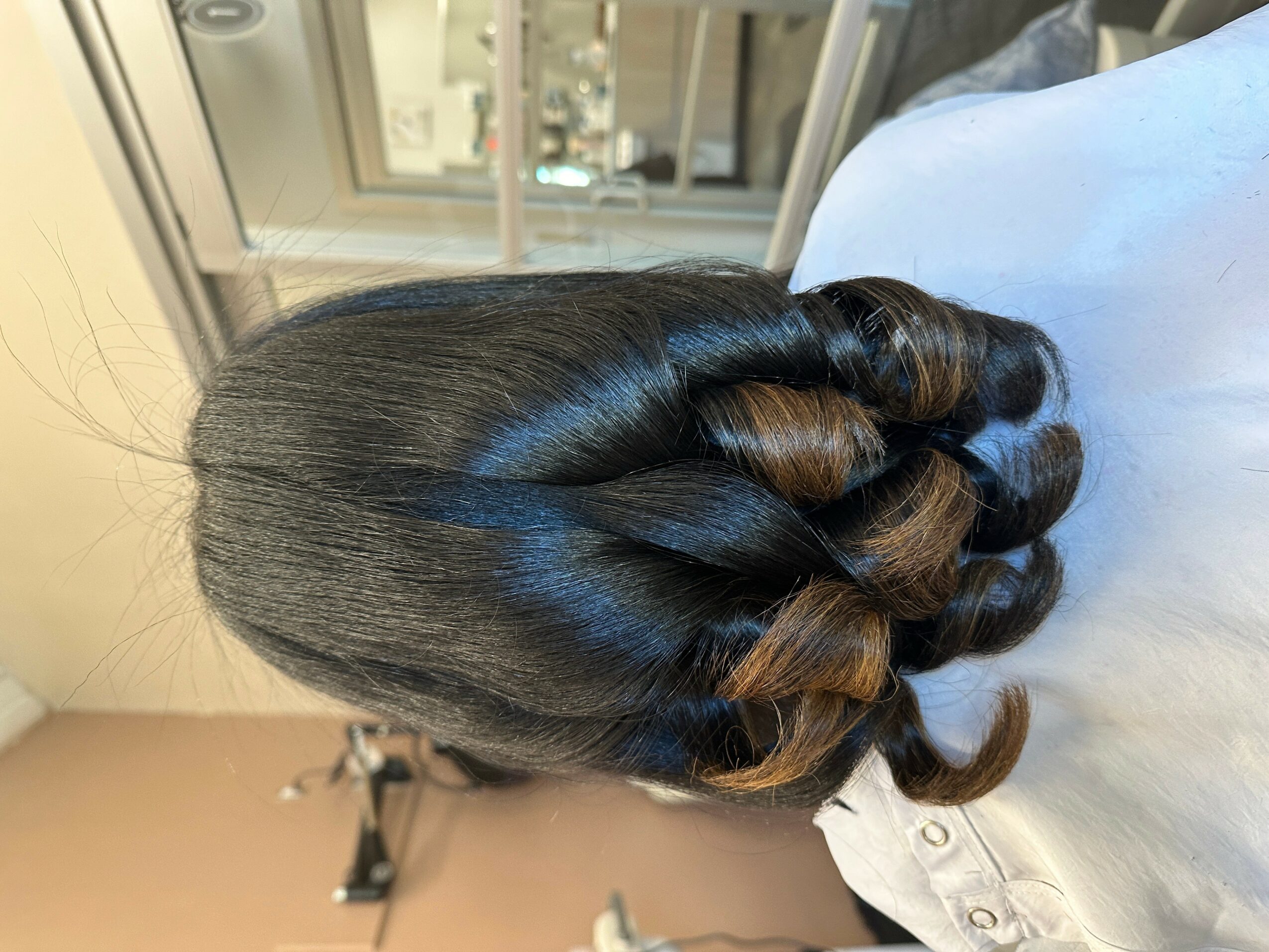 Side view of a person with styled, shiny black and brown hair in a twisted updo, sitting indoors near a window.