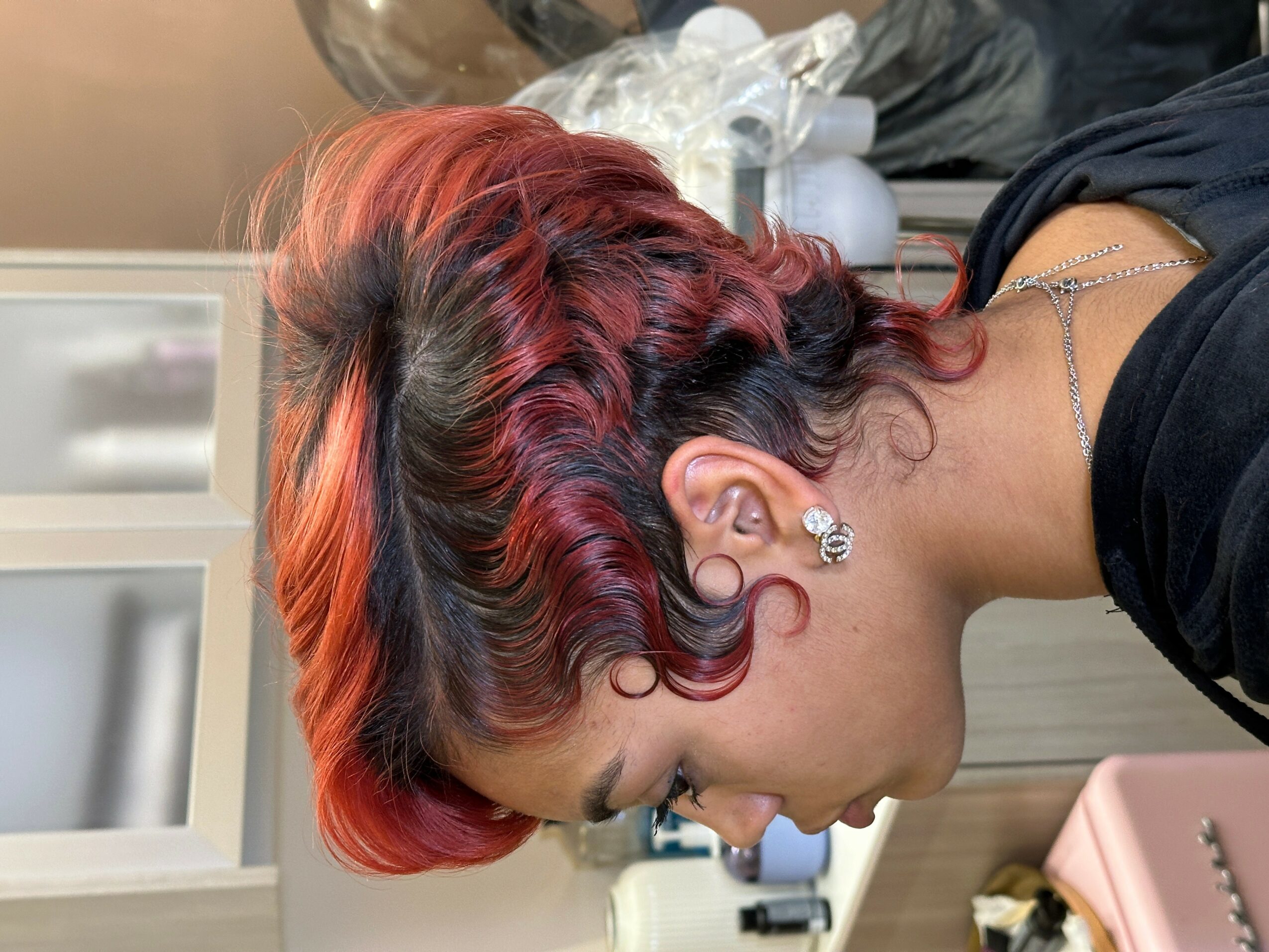Side view of a woman with red and black curly hair, wearing earrings and a black top, in a room with cabinets.
