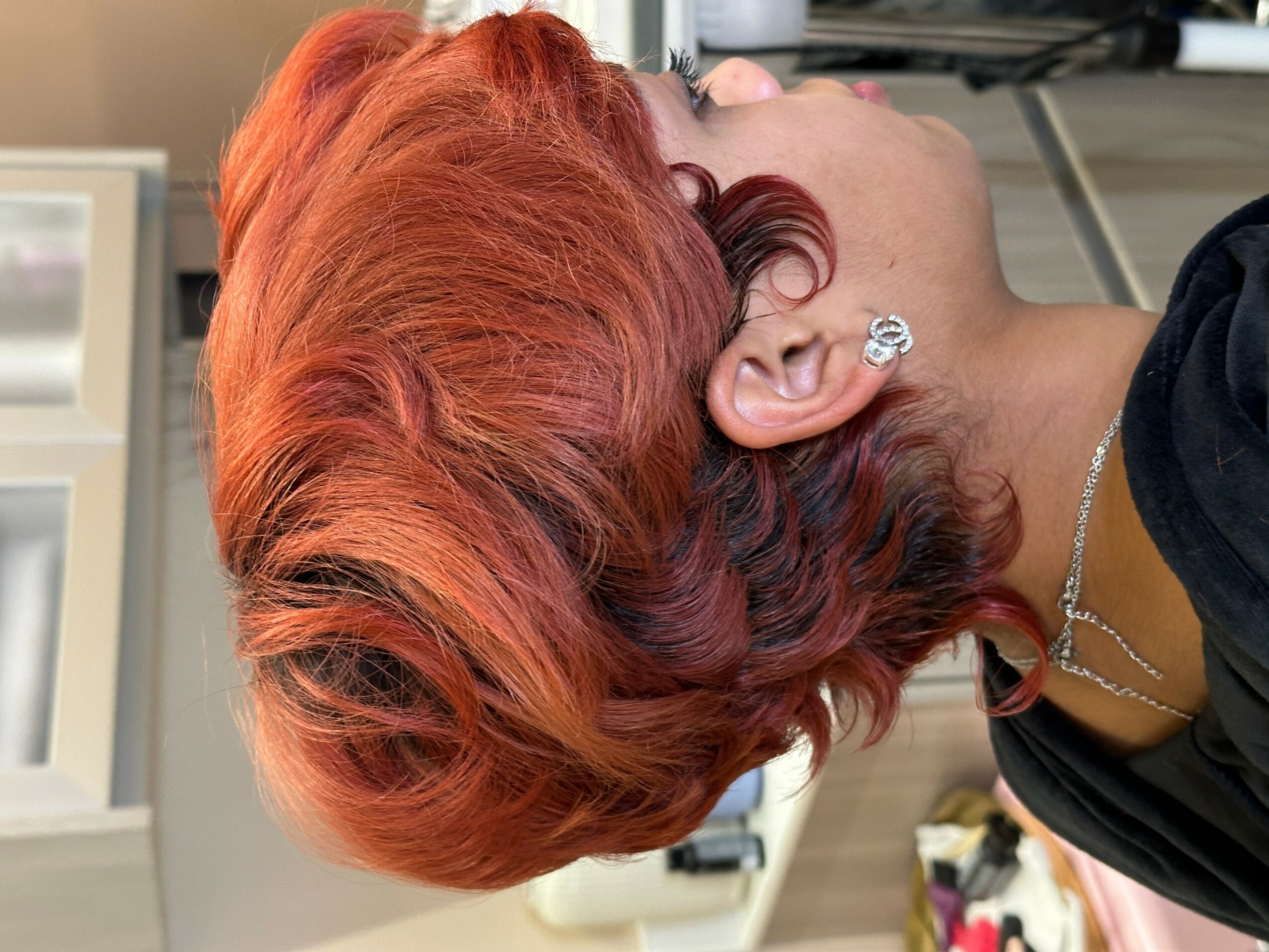 Side view of a woman with vibrant red and dark hair, wearing earrings and a necklace, indoors.