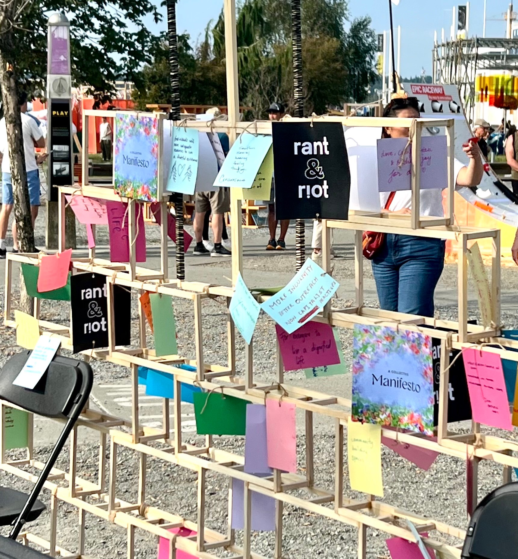 Wooden frame with colorful notes and signs, people walking in background, outdoor setting with trees and buildings.