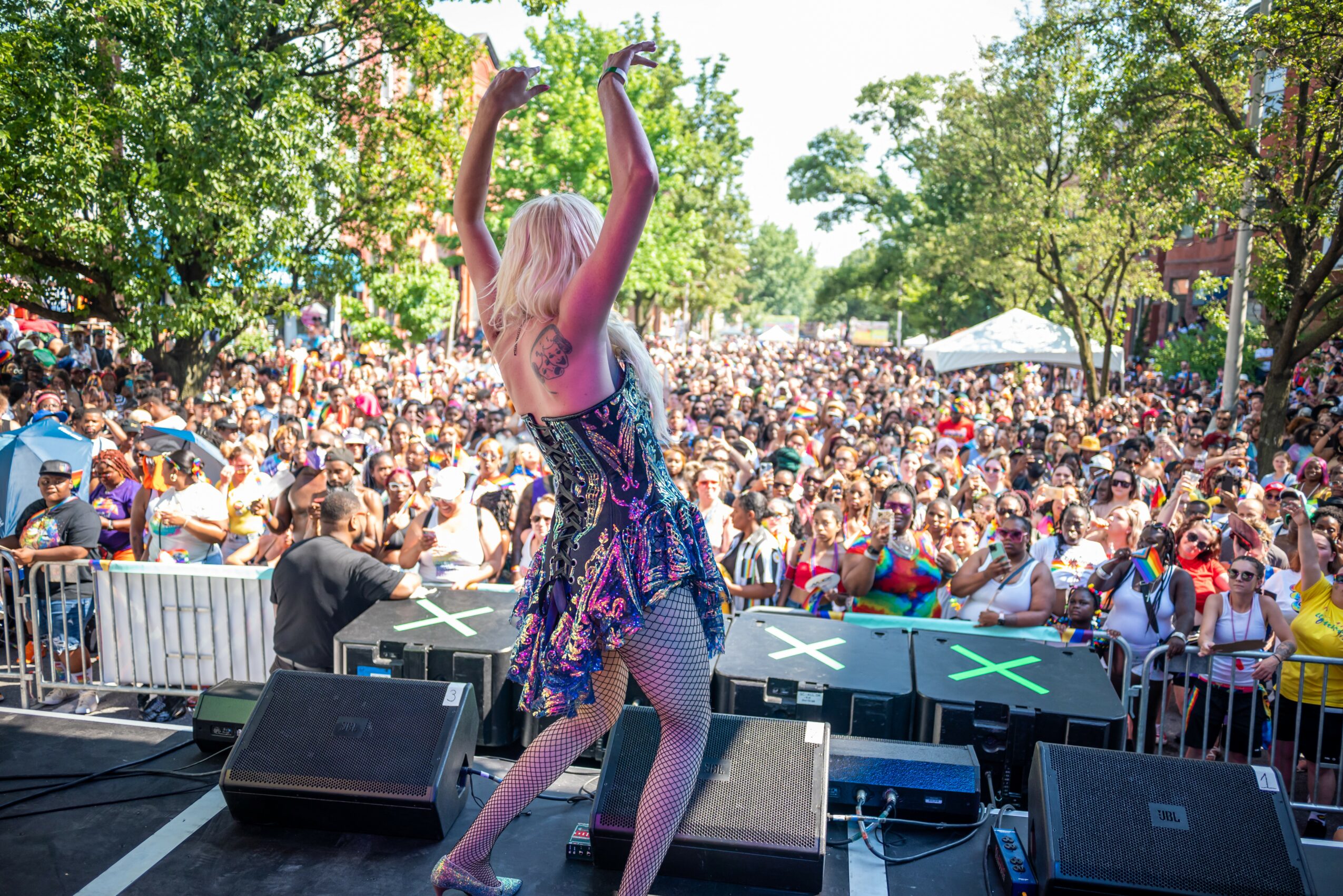 Performer with long blonde hair dancing on stage in front of a large crowd outdoors during daytime.