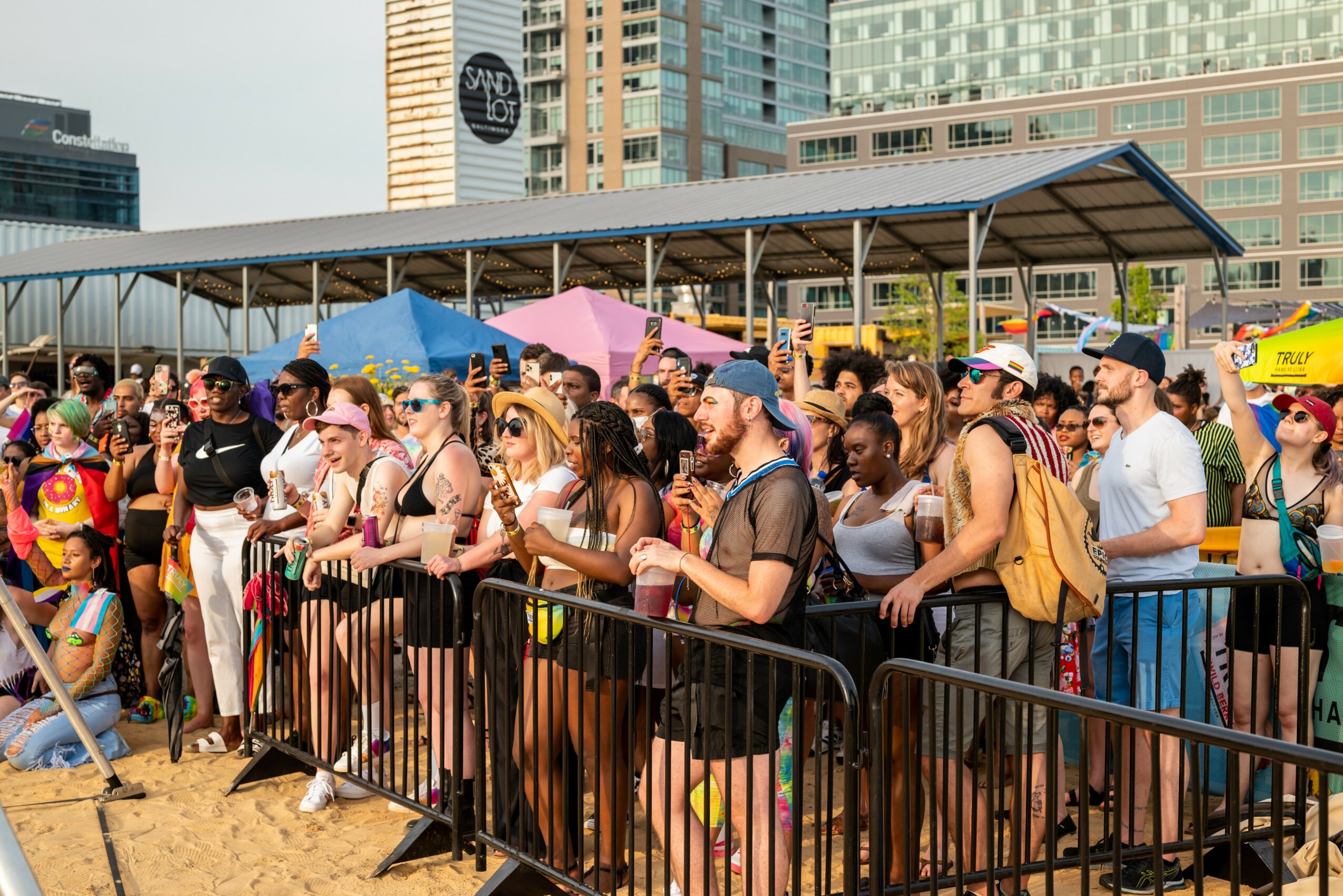Crowd of people waiting behind barriers outdoors with tall buildings in background.