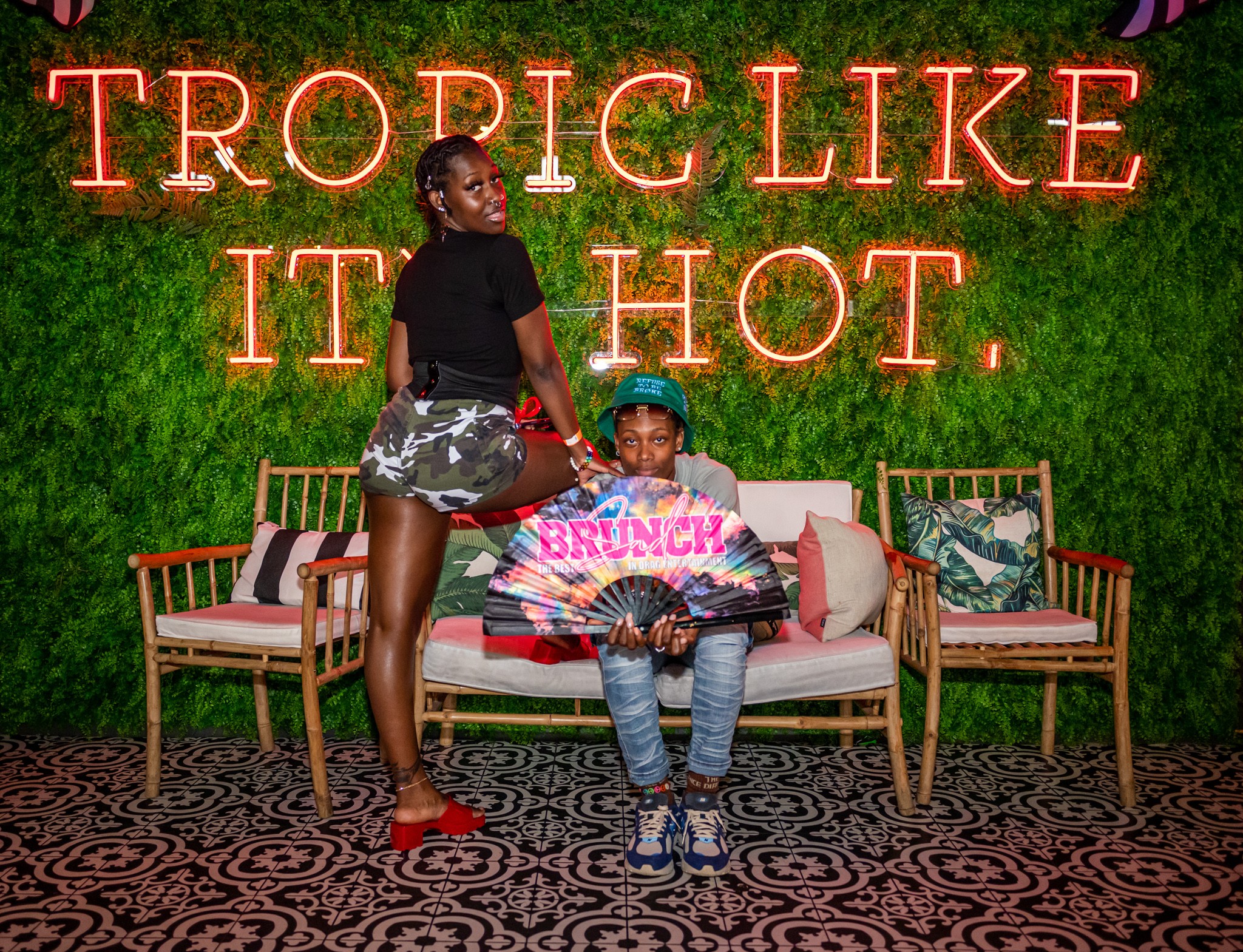 Two people in front of a neon sign that reads 'Tropic Like It Hot,' with a green wall and patterned floor.