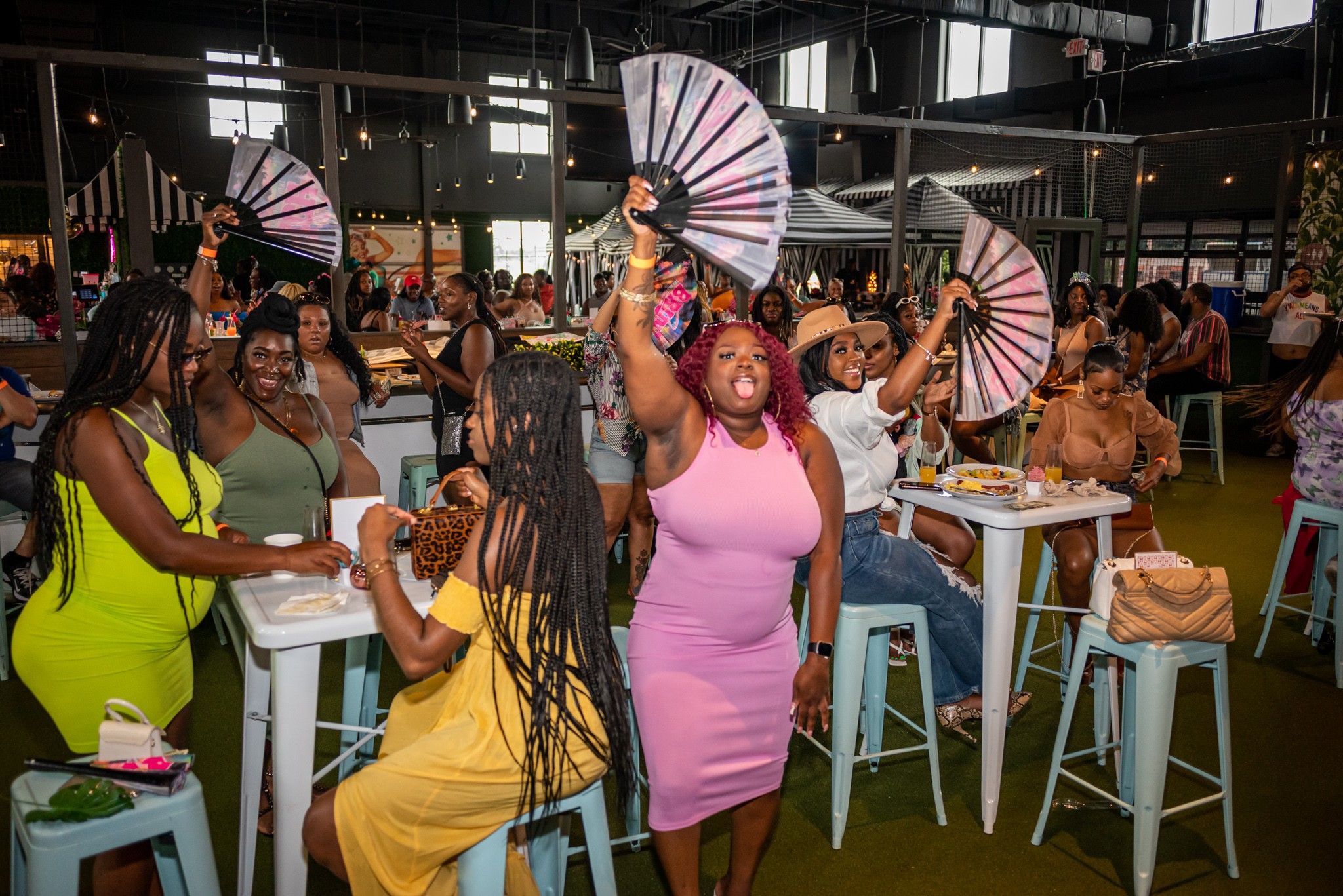 Group of women dancing and holding fans at a lively indoor event with tables and chairs.