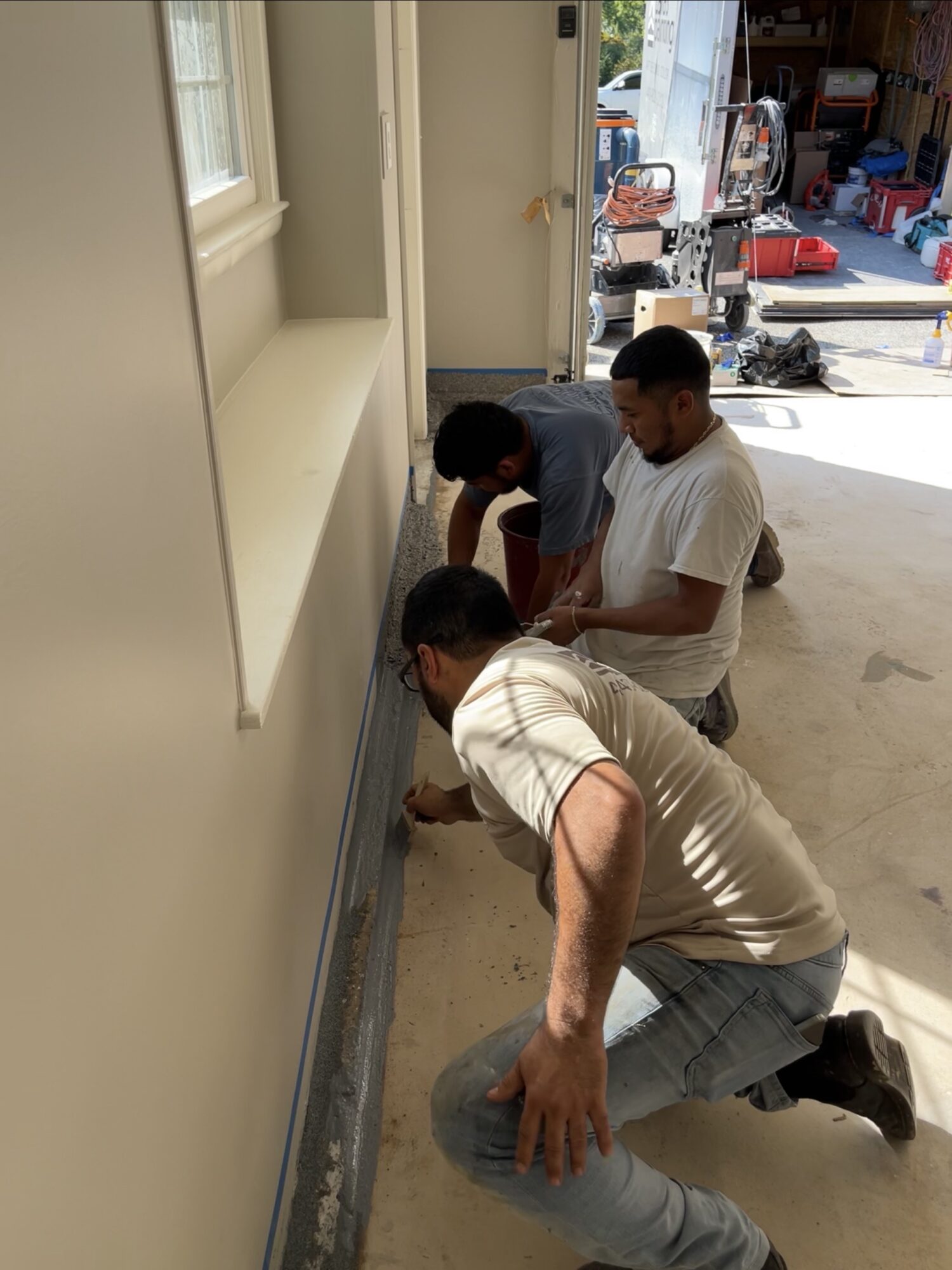 Three people working on a baseboard in a garage, with tools and storage in the background.