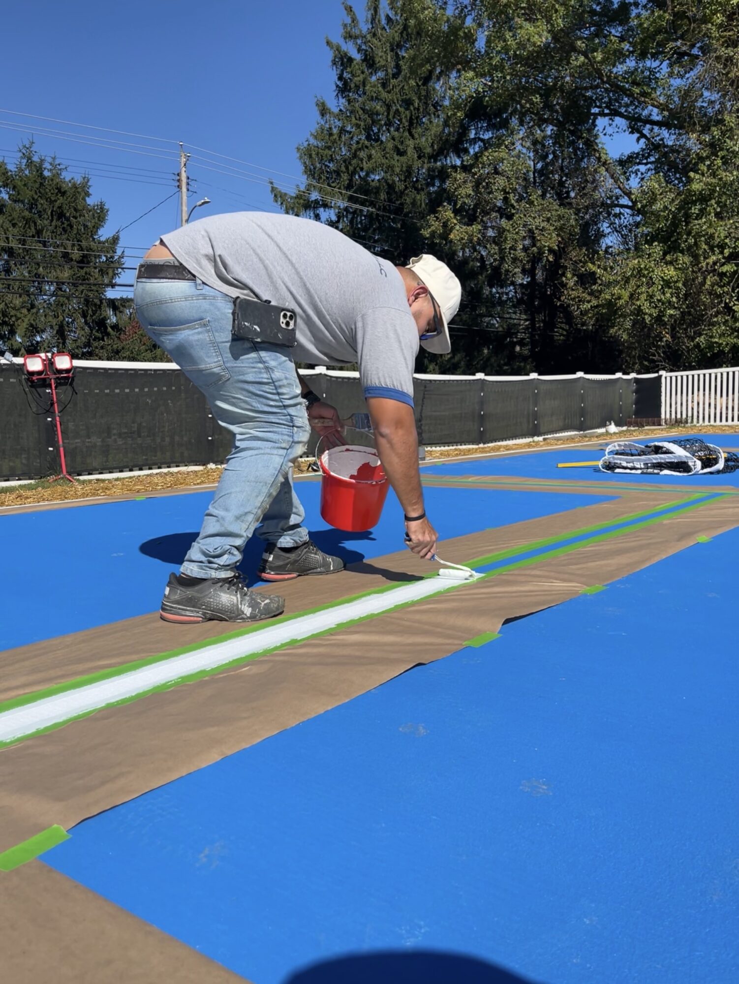 Man painting a line on a surface outdoors with trees and a fence in the background.