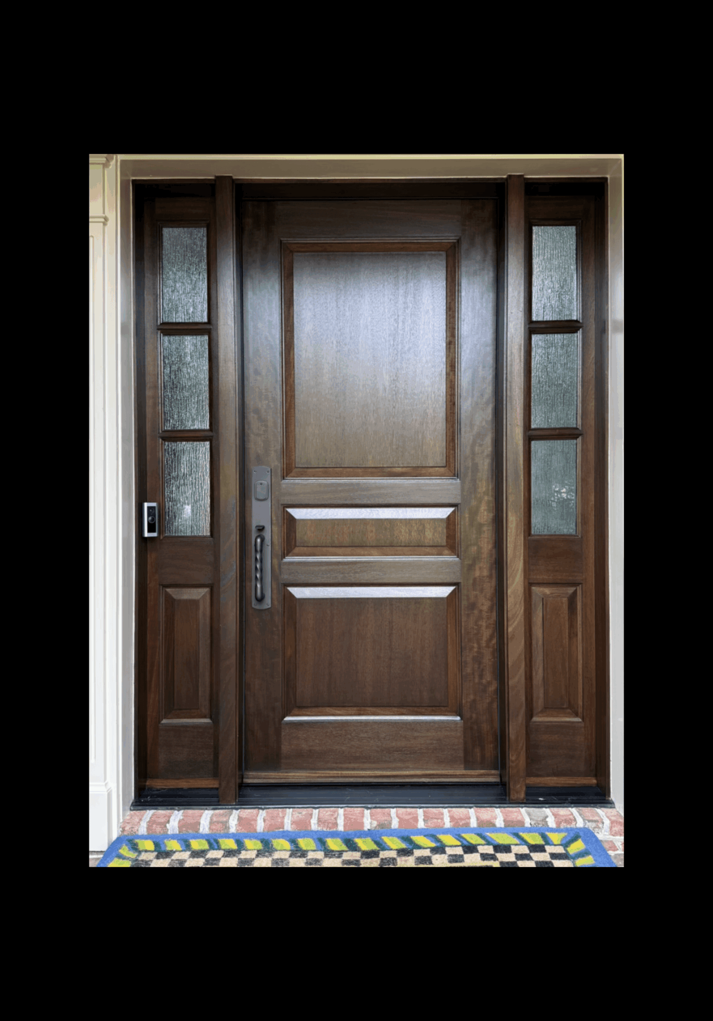 Wooden front door with glass panels on sides, decorative panels, and a colorful tiled threshold.