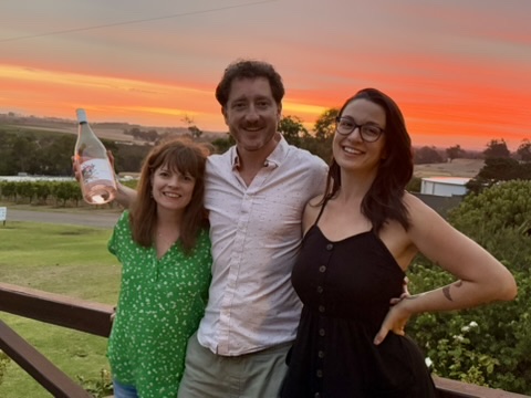 Three people smiling outdoors at sunset, with a woman holding a bottle, a man in the middle, and a woman on the right, all standing on a wooden railing.