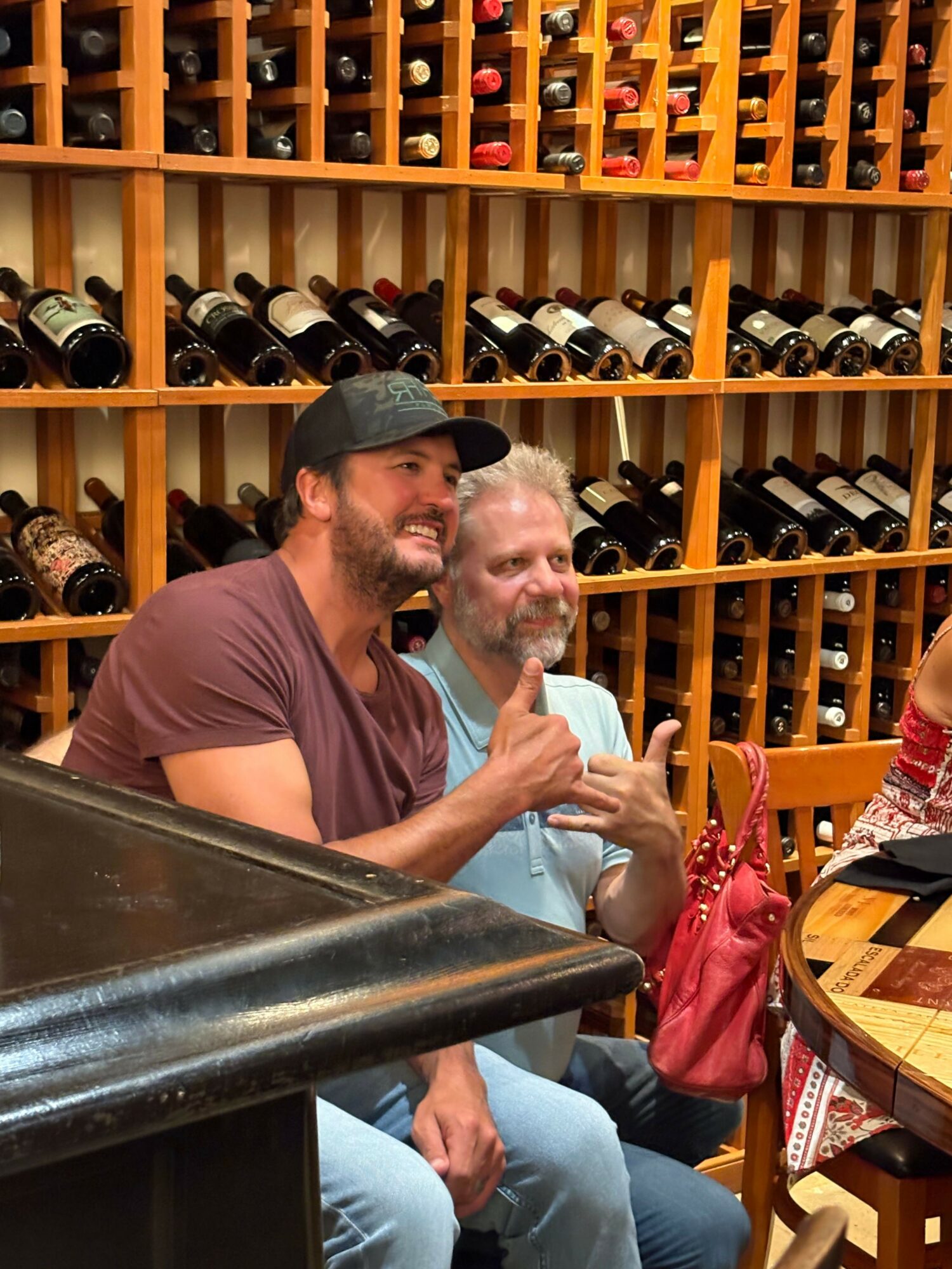 Two men sitting at a bar counter, one with a beard and the other with a cap, in a wine cellar setting.