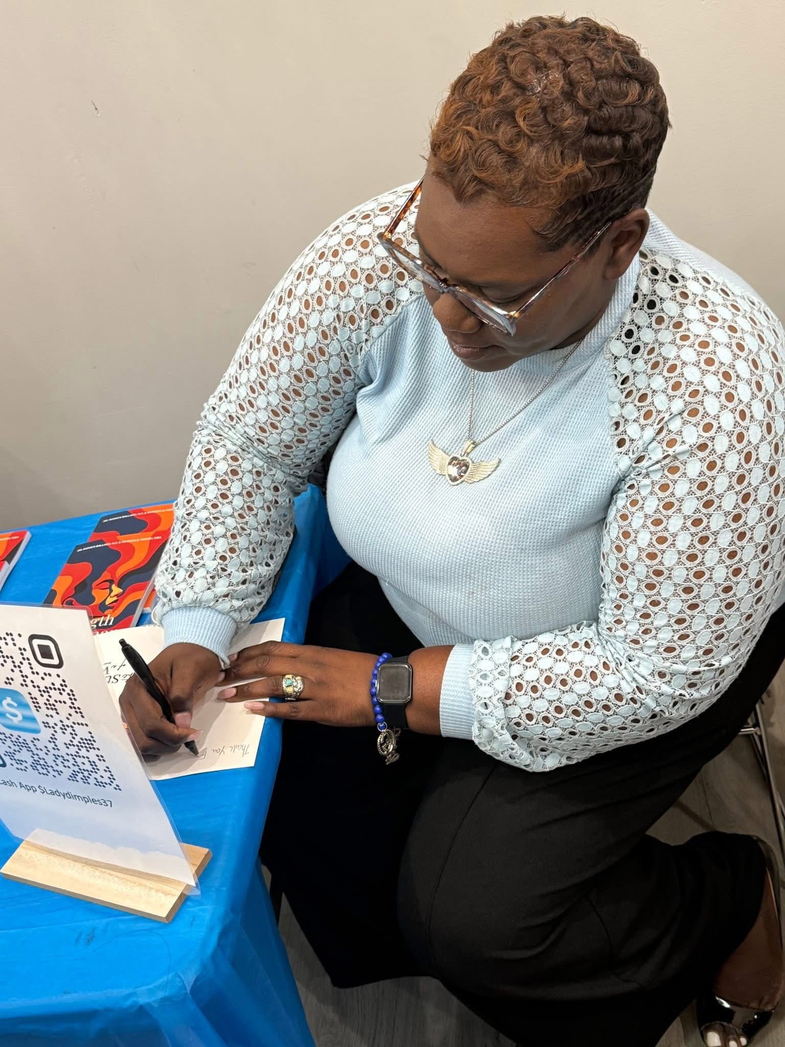 Woman with short curly hair and glasses writing at a table with a blue tablecloth, QR code, and colorful book, viewed from above.