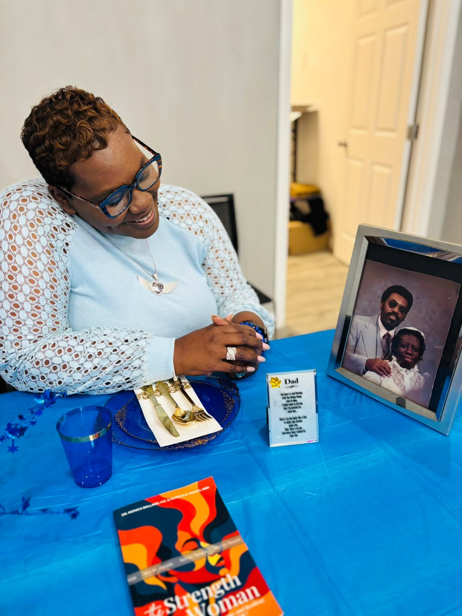 Woman with short curly hair and glasses smiling at a laptop during a virtual meeting, sitting at a blue table.