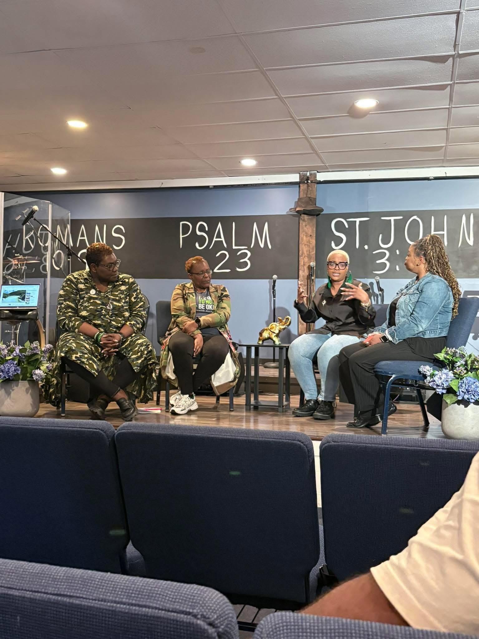 Four people seated on a stage in a church setting, with a blackboard behind them displaying 'PSALM 23' and 'ST. JOHN 3'.