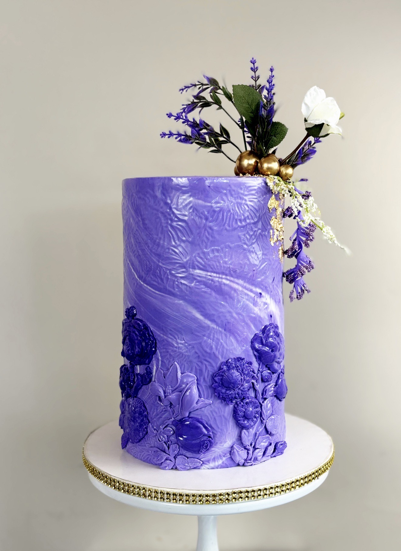 Tall purple cake with floral decorations on top, placed on a white stand with gold accents.