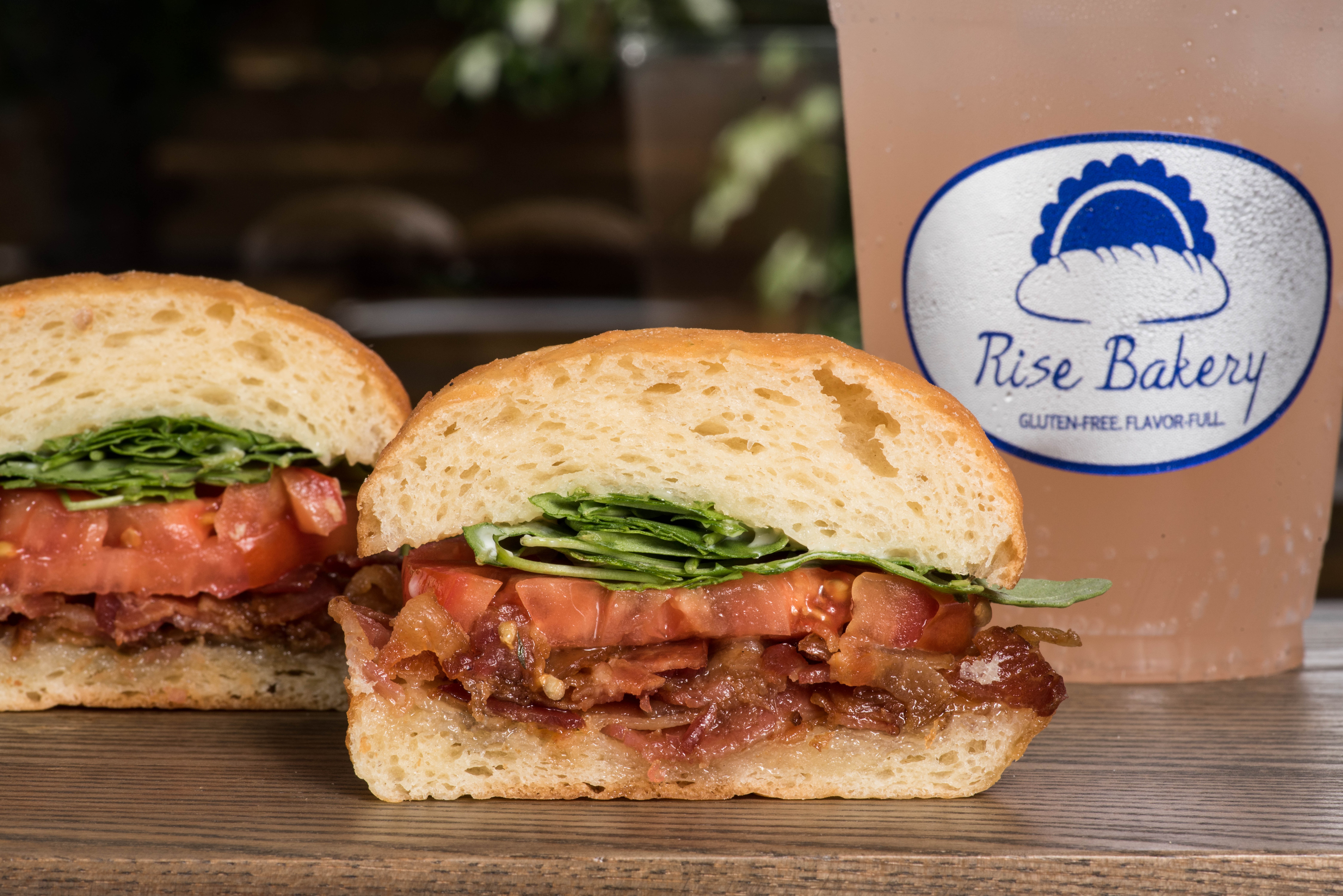 Sandwich with tomato, lettuce, and meat, next to a drink with Rise Bakery logo, on a wooden surface.