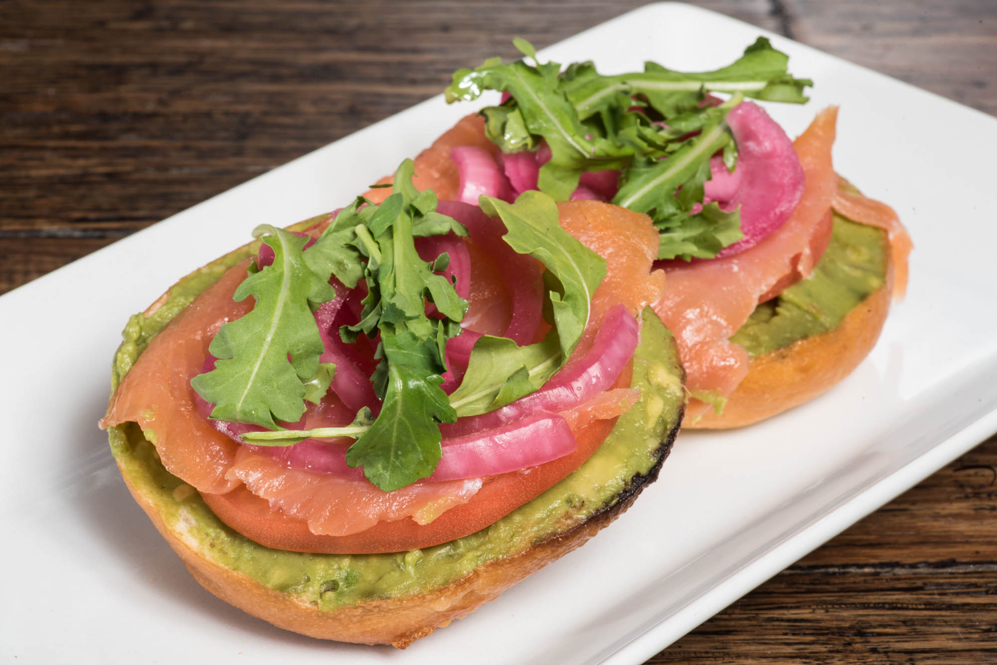 Two avocado toast slices topped with smoked salmon, radish slices, and mixed greens on a white plate.